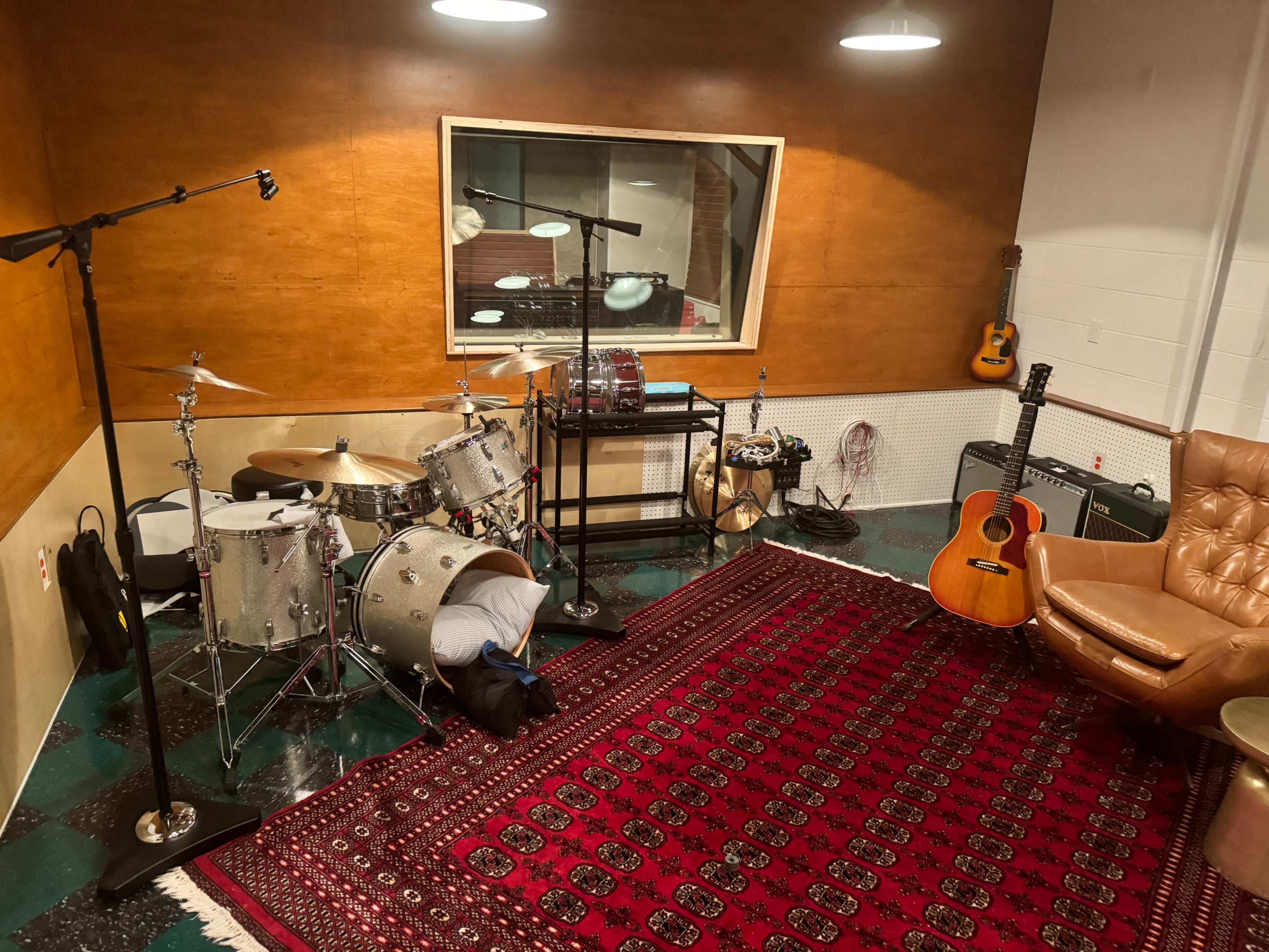 A music studio features a drum set, an acoustic guitar, microphones on stands, and a large rug in a room with wooden walls and studio equipment visible through a window.