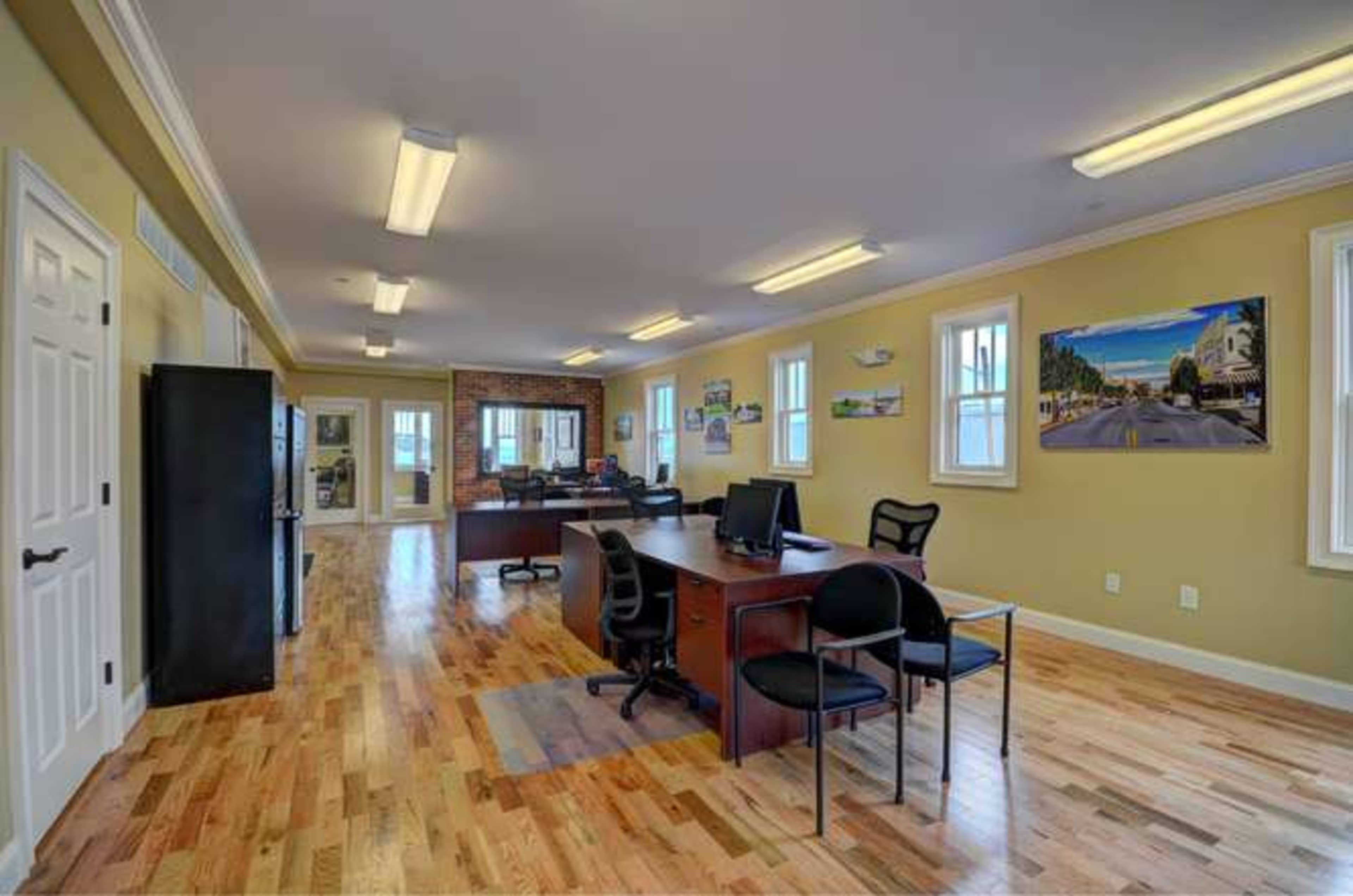 The image shows a spacious office with wooden floors, desks, and computers, featuring large windows and artwork on the walls.