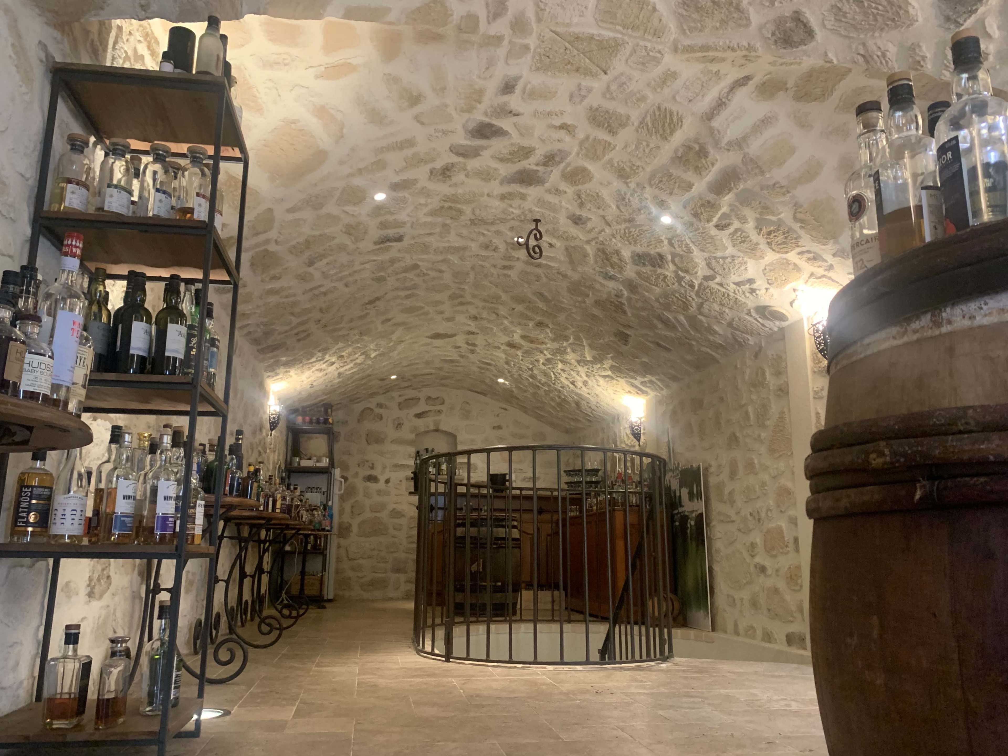 A stone-walled cellar with shelves of liquor bottles, wooden barrels, and a circular bar area in the center.