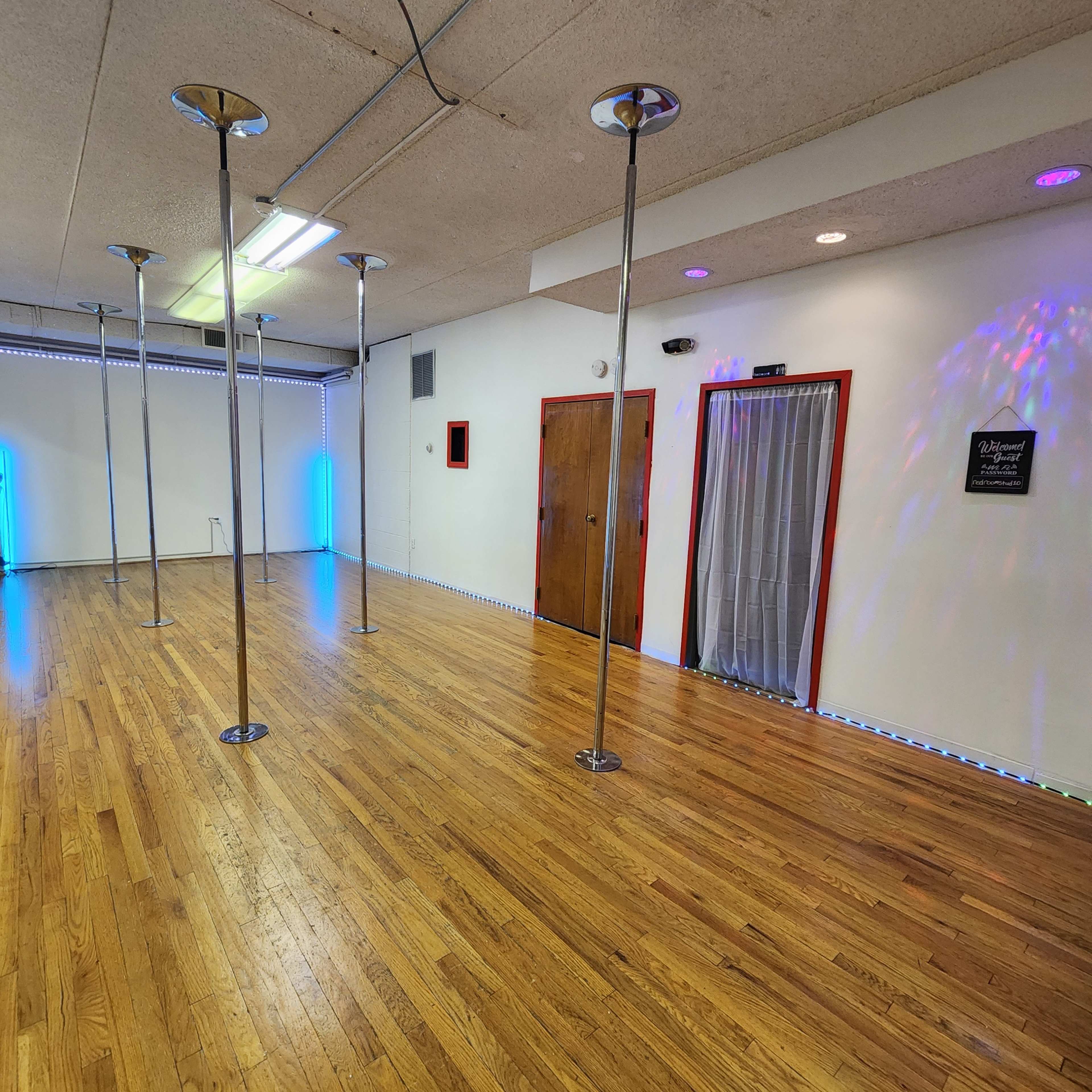 The image shows an empty wooden-floored room with several shiny metal poles and colorful lighting along the walls.
