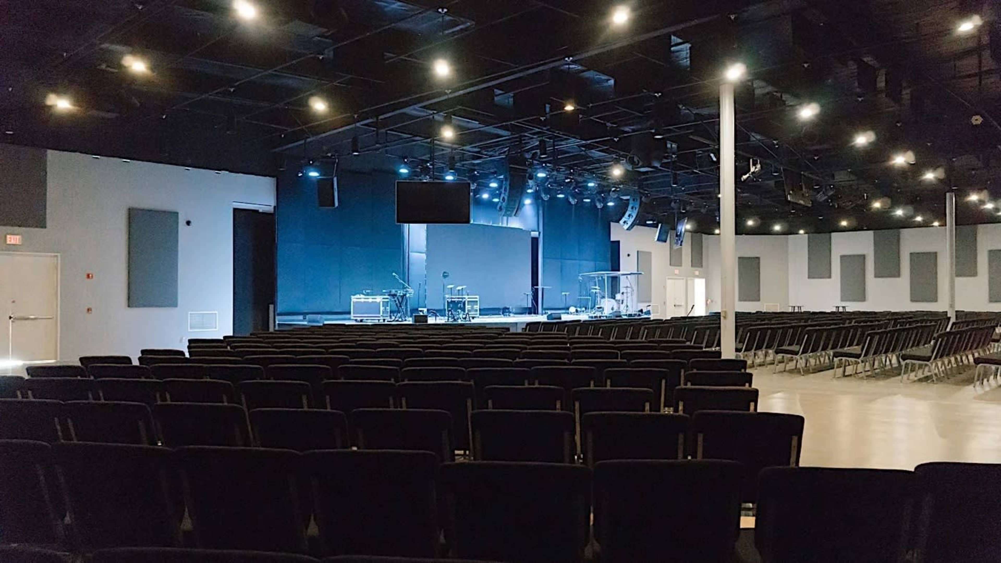 An empty auditorium with rows of black chairs facing a stage equipped with sound and lighting equipment.