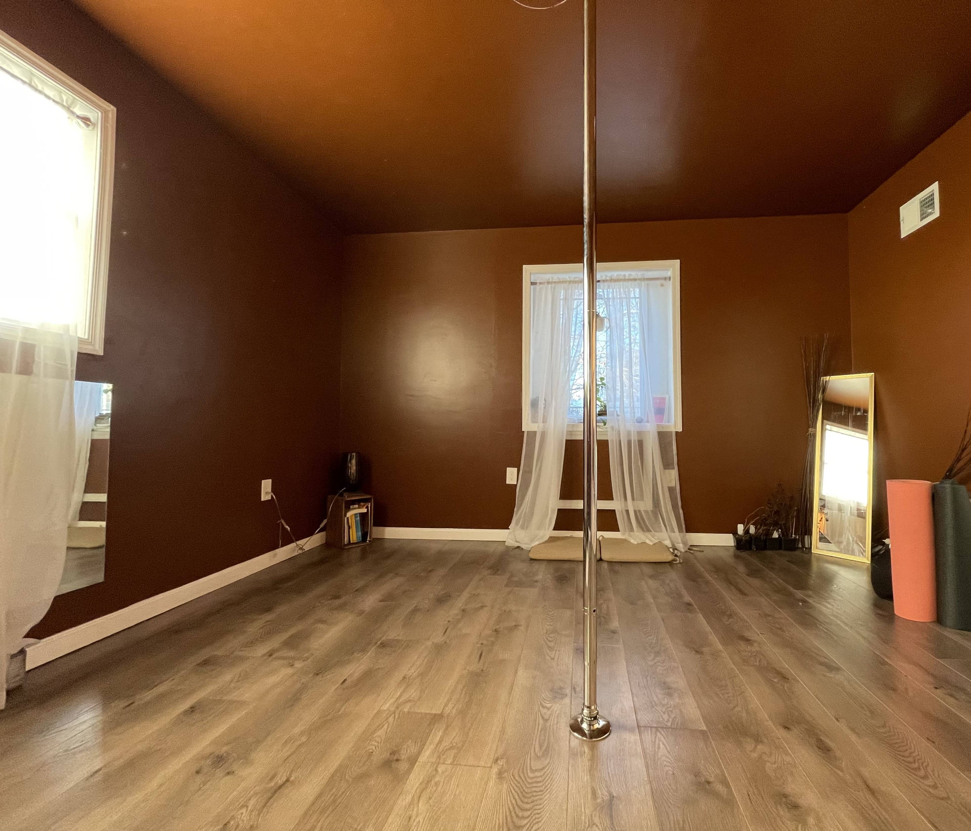 A spacious room with brown walls, wooden flooring, a pole in the center, and large windows adorned with sheer curtains.