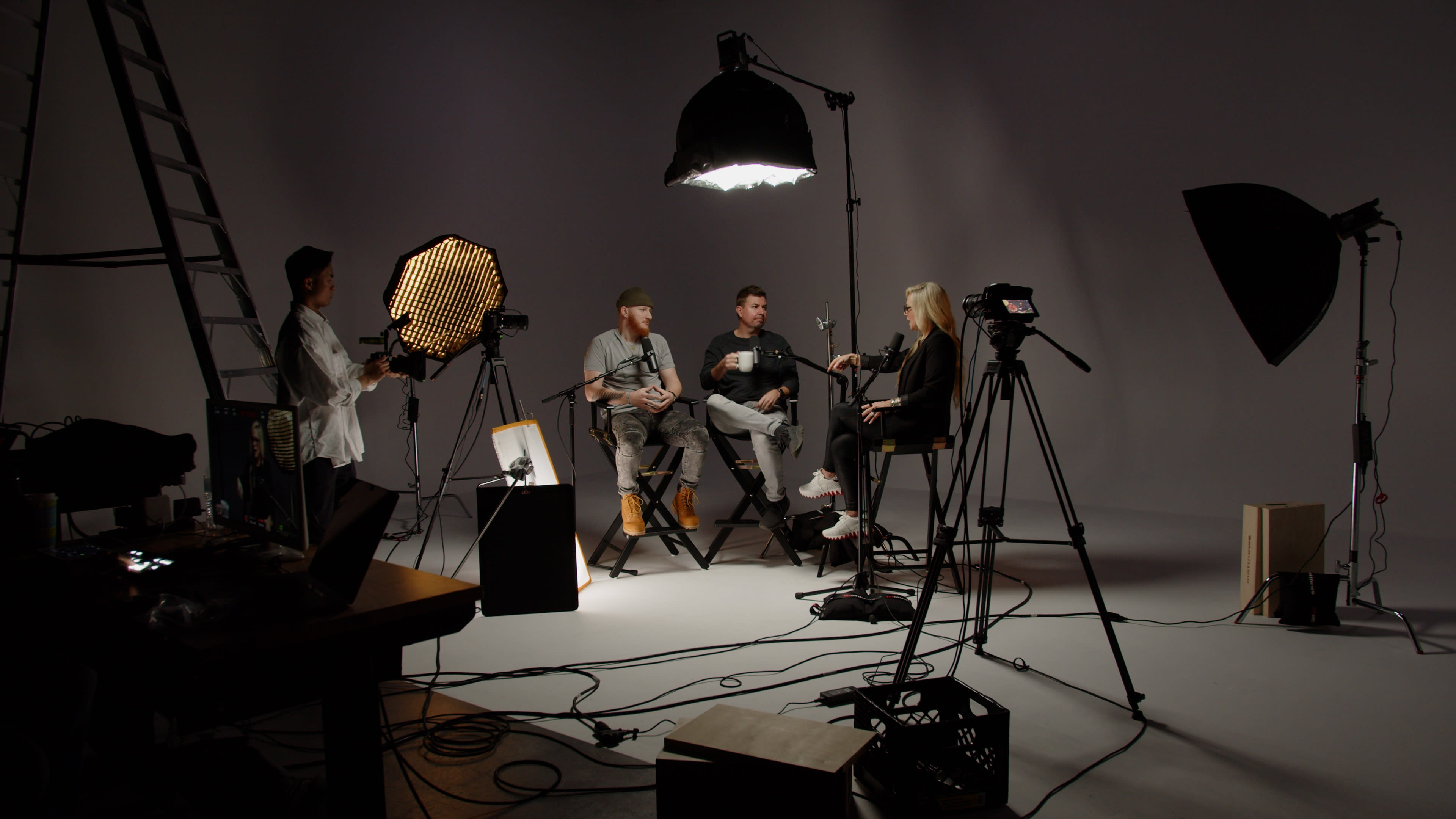 A video shoot is set up in a studio with three people seated on director's chairs, surrounded by various lighting equipment and cameras.