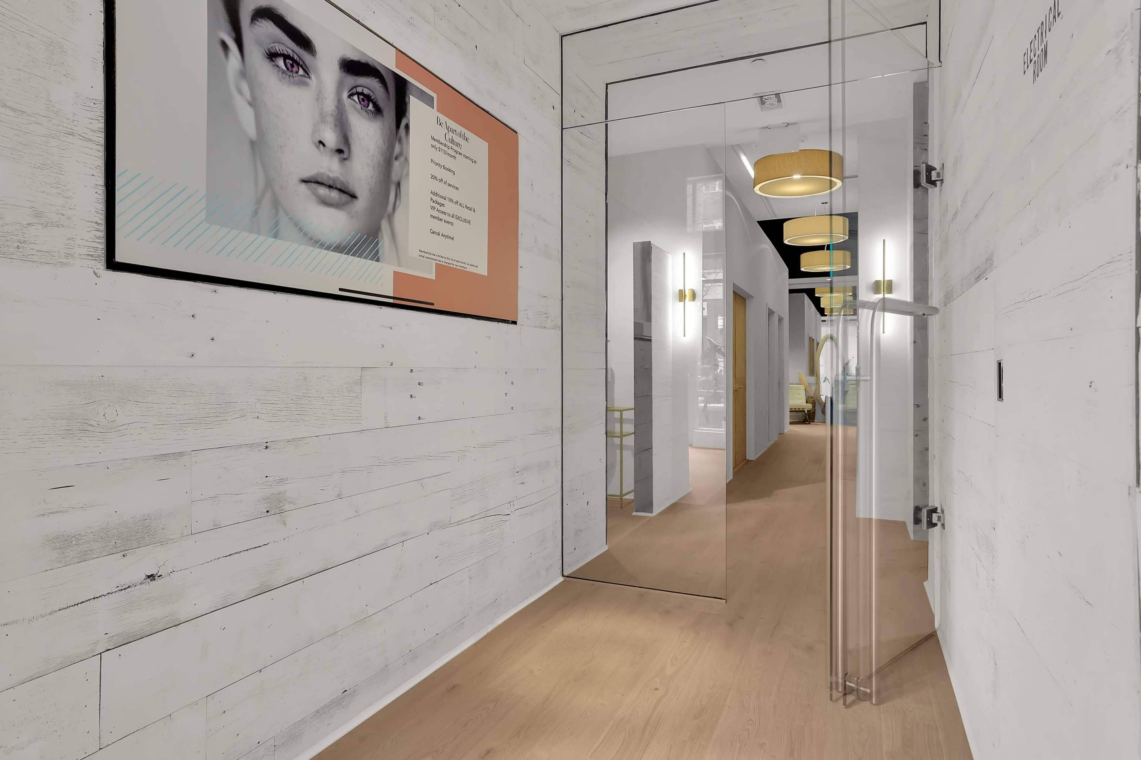 A modern hallway features a large portrait on a wall, leading to a bright area with pendant lights and glass doors.