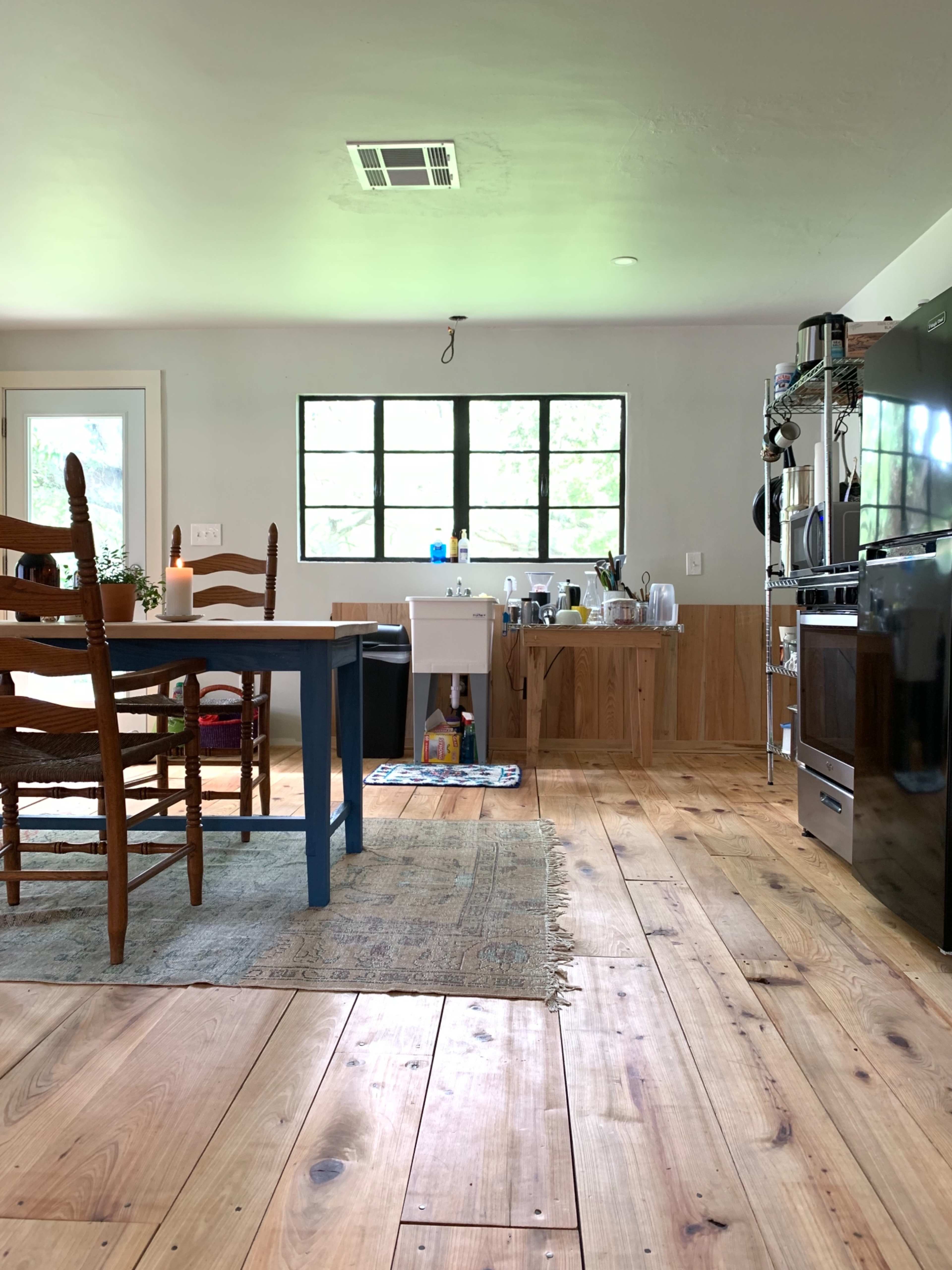 A kitchen features a wooden table and chairs, a black refrigerator, and a sink with dishware on the counter, all set against a backdrop of wooden flooring and a window.