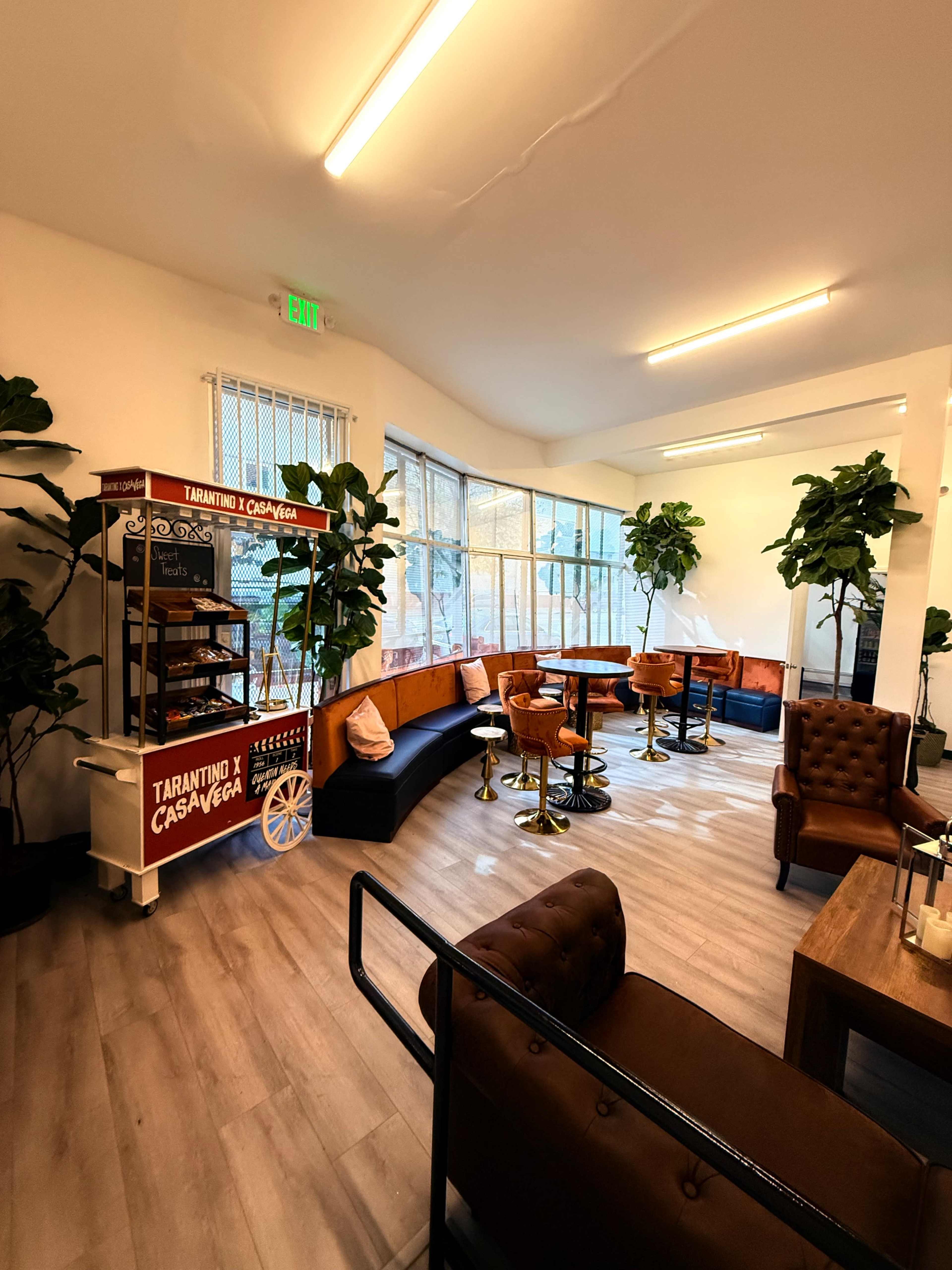 The image shows a modern lounge area with brown leather seating, tall tables, and green potted plants, alongside a vintage-style cart labeled "Tarantino X Casa Vega."