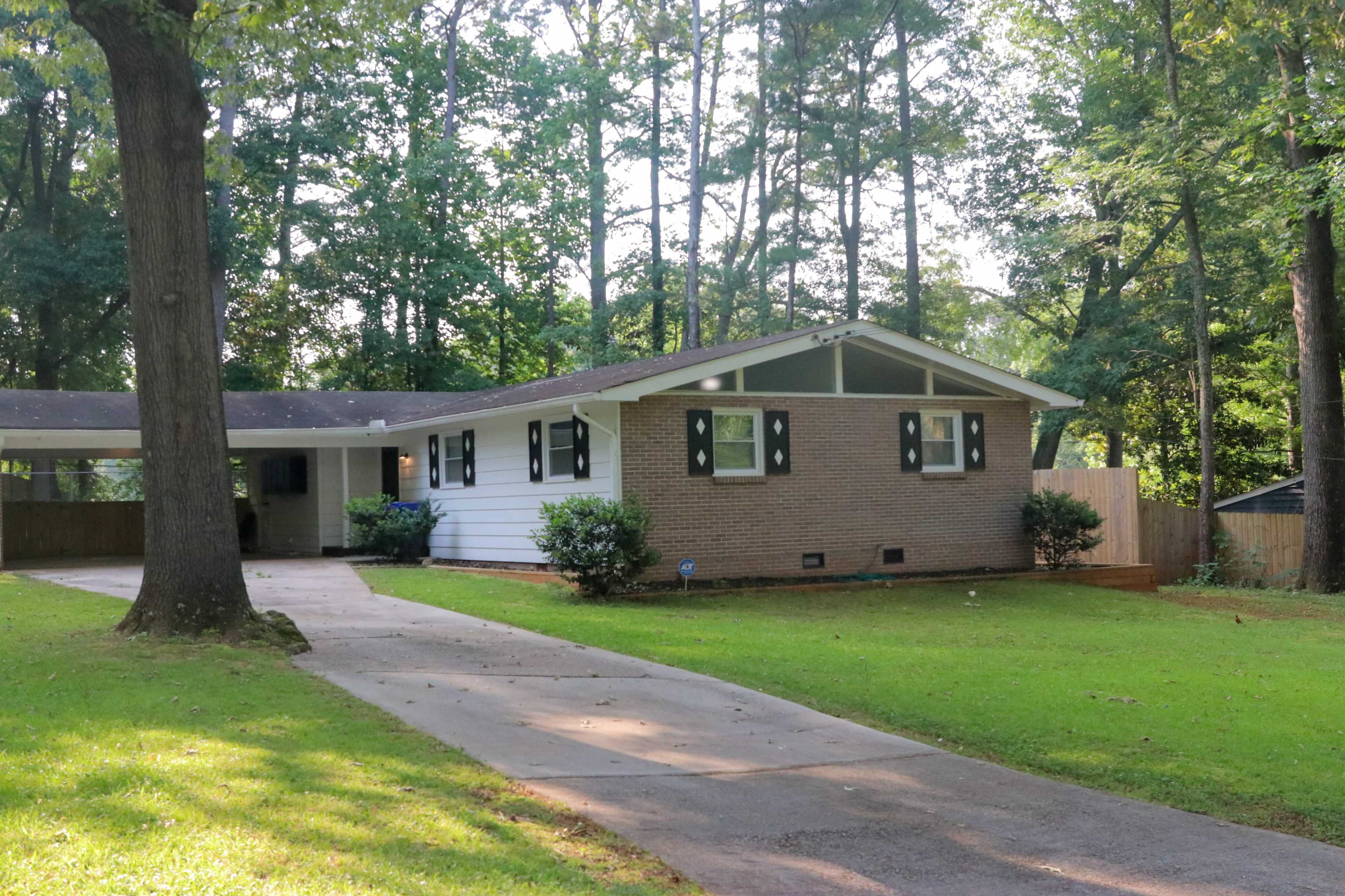 The image shows a single-story house with a brick front and white siding, situated on a lawn with trees surrounding it, along a paved driveway.