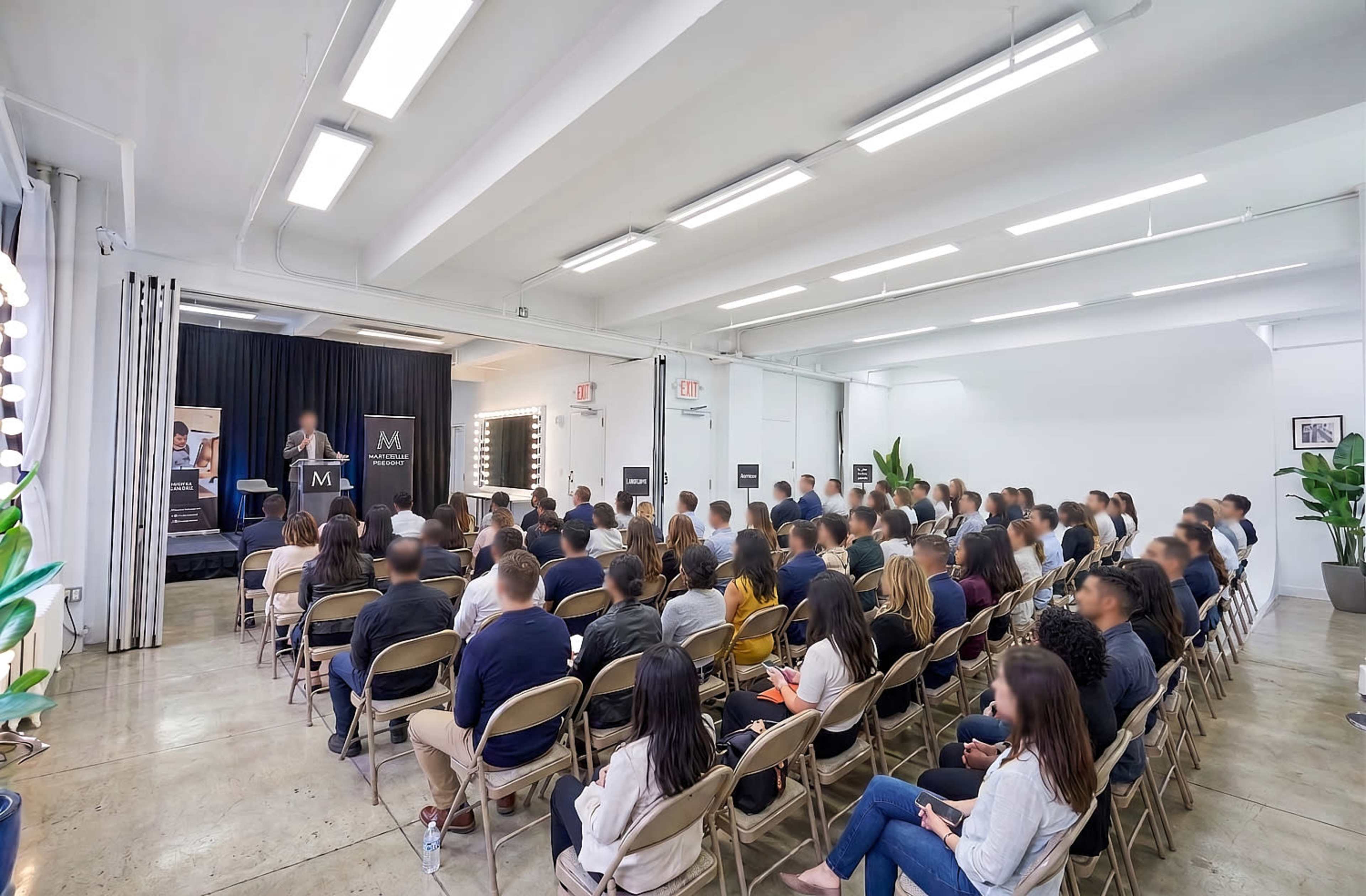 A large audience sits in foldable chairs, facing a speaker on a stage in a well-lit, modern venue.