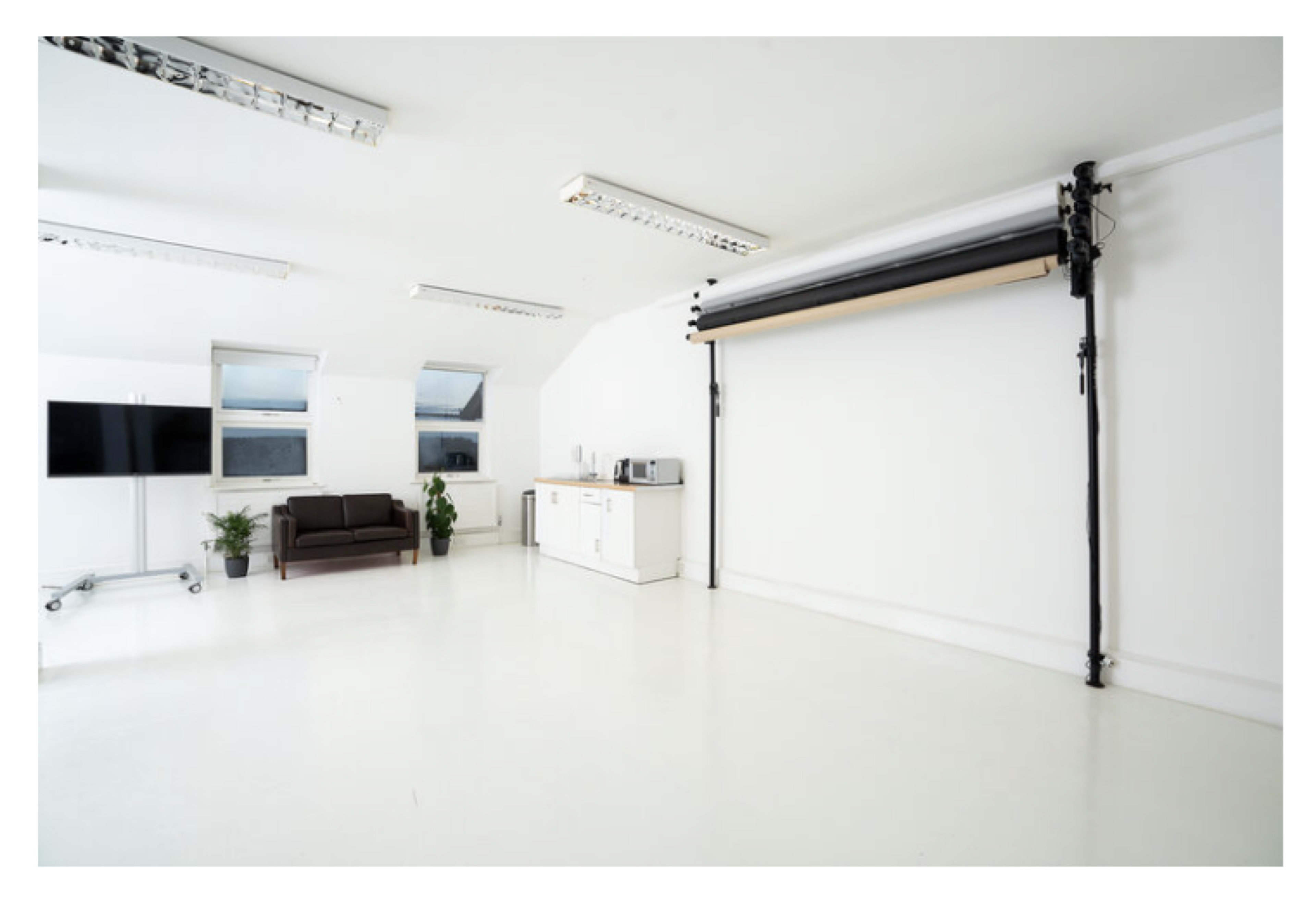 The image shows a spacious, well-lit room featuring a couch, a kitchenette with appliances, and a roll-down backdrop for photography.