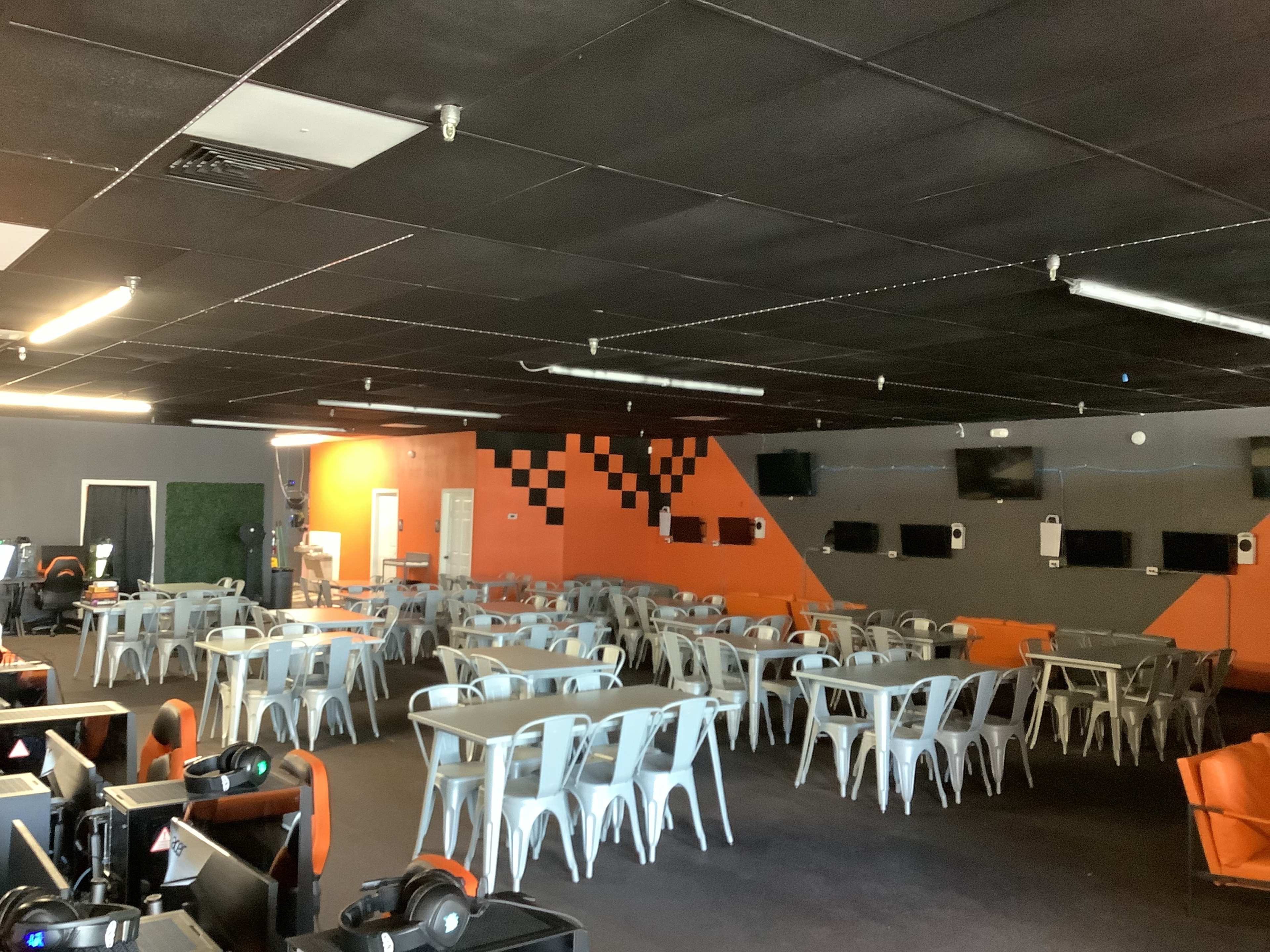 The image shows a spacious gaming room with multiple rows of metallic chairs and tables, orange and gray walls, and several screens mounted on the walls.
