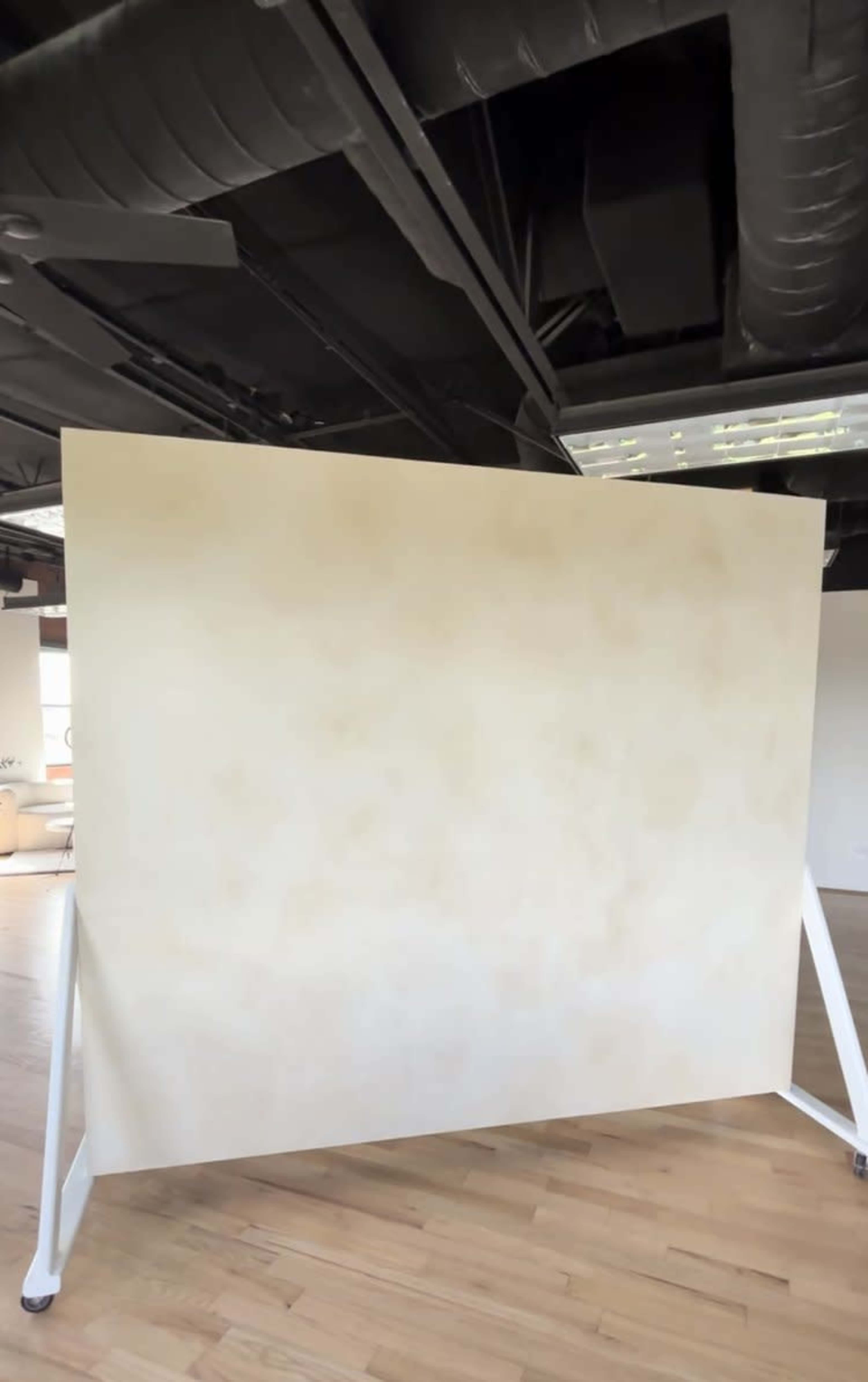 A large, blank canvas on a freestanding frame is positioned in a room with wooden flooring and exposed black ceiling beams.