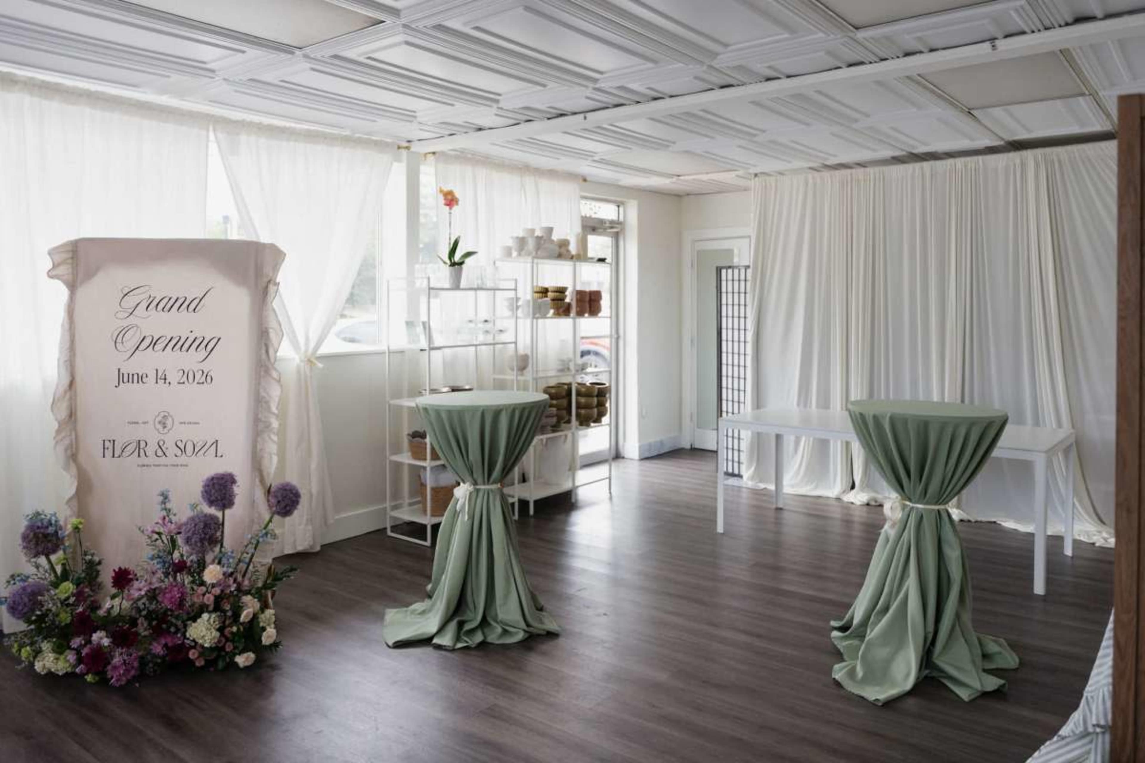 The image shows a decorated interior space for a grand opening, featuring tables draped in green fabric, floral arrangements, and a backdrop with event details.