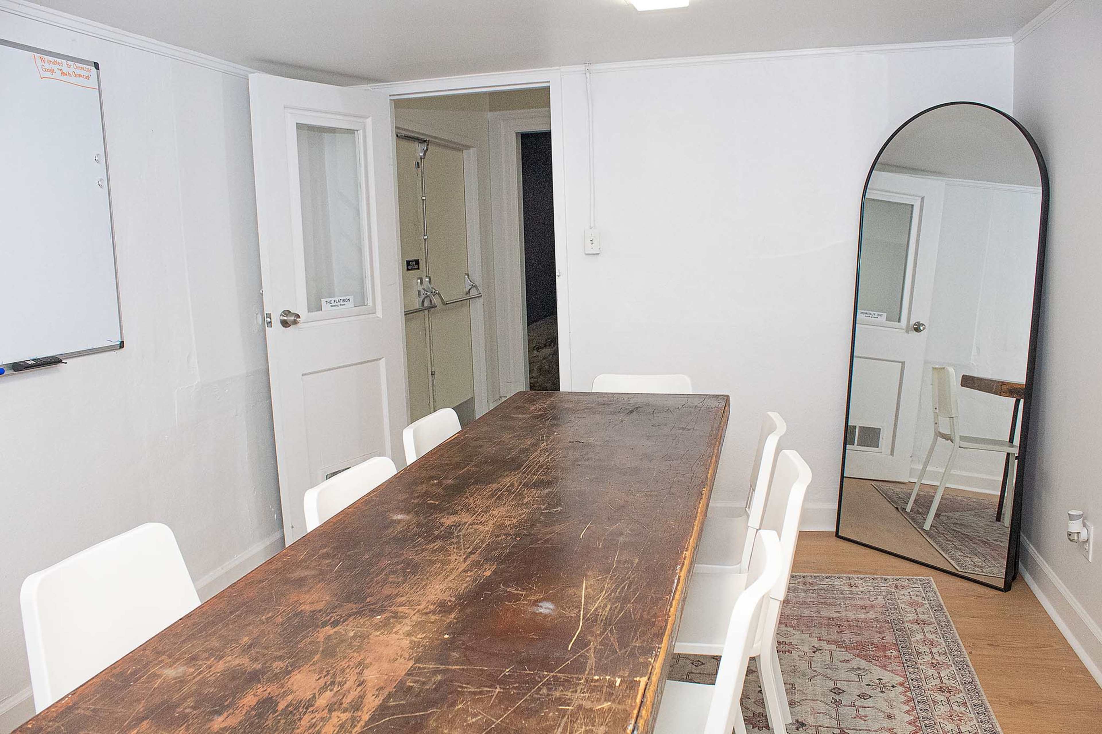 A simple meeting room with a wooden table surrounded by white chairs, a large mirror against the wall, and a whiteboard mounted on the wall.