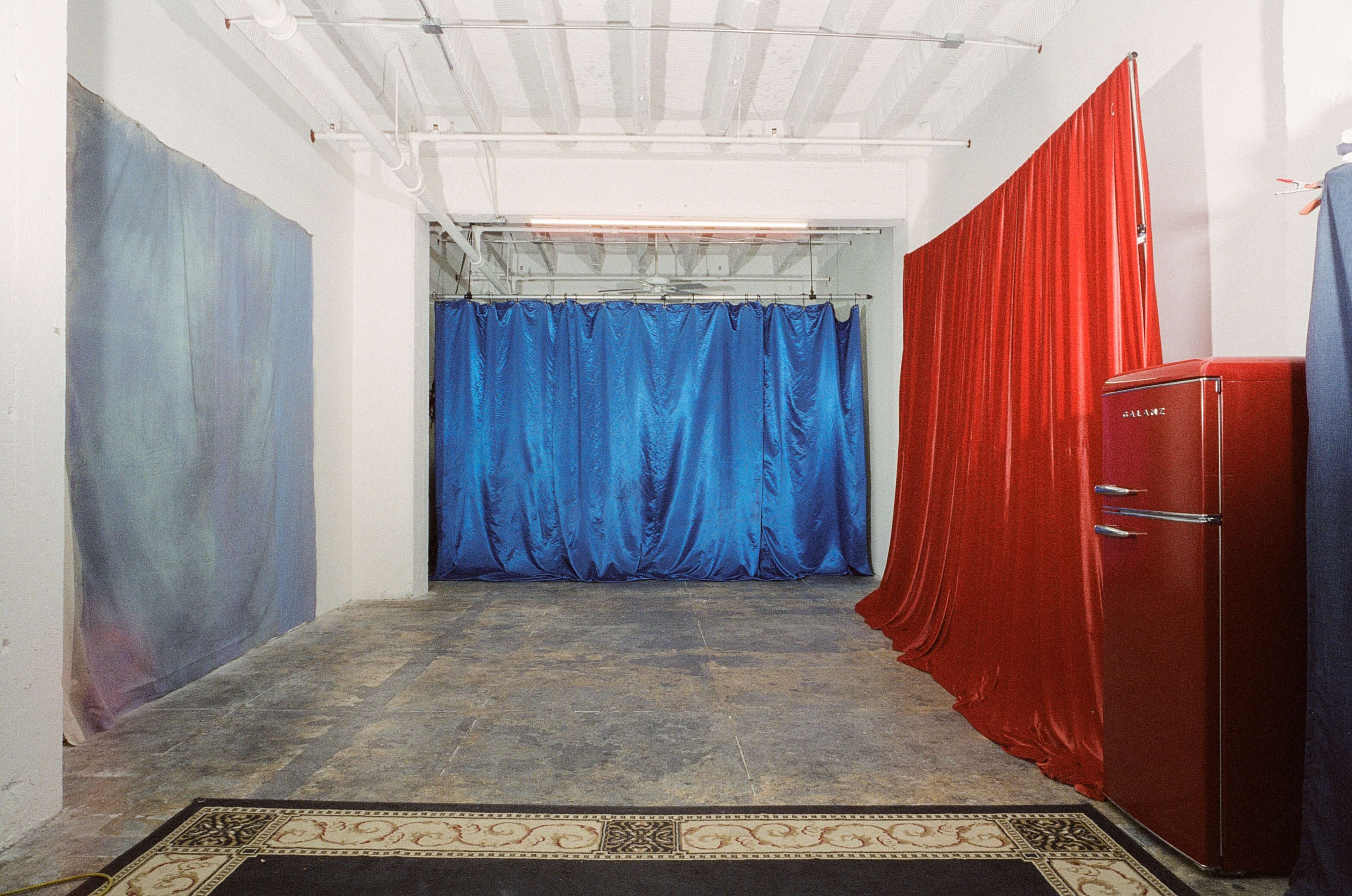 The scene shows a room with a concrete floor, draped red and blue curtains on the walls, and a vintage red refrigerator positioned in the corner.