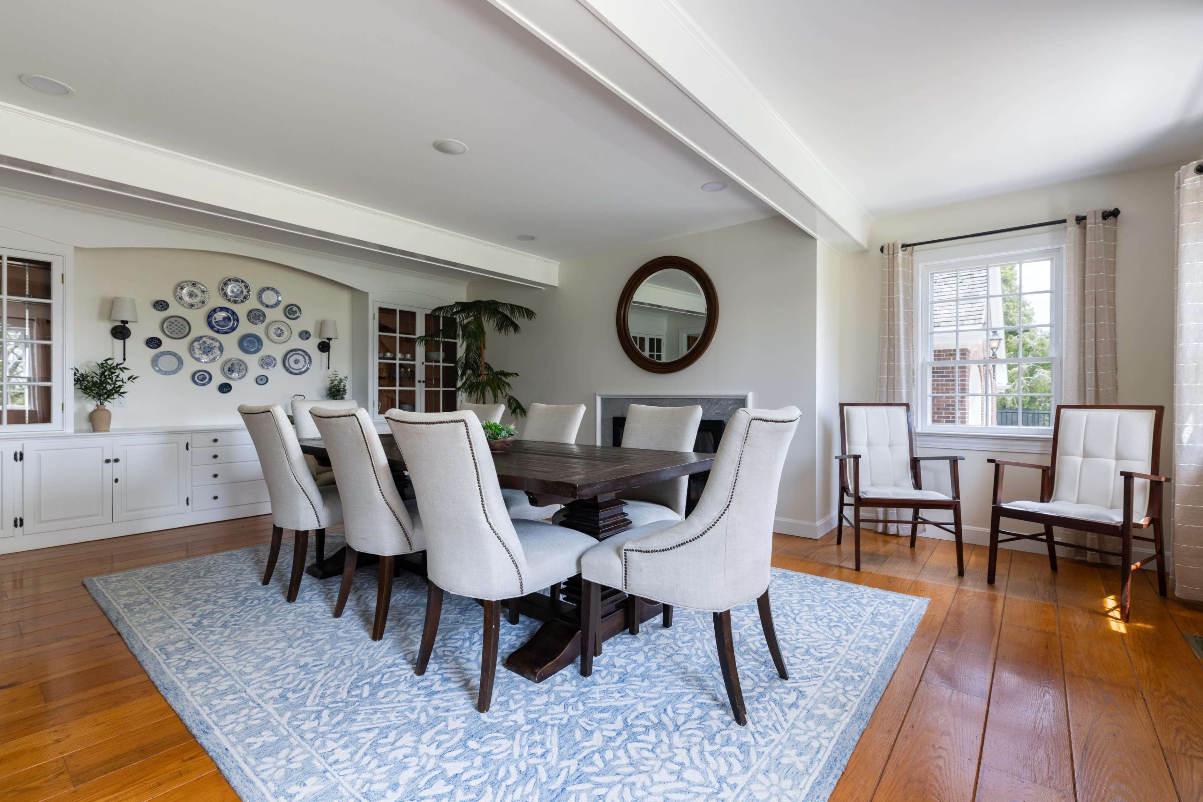 The image shows a dining room with a large wooden table surrounded by upholstered chairs, a display of decorative plates on the wall, and additional seating in the form of two armchairs near the windows.