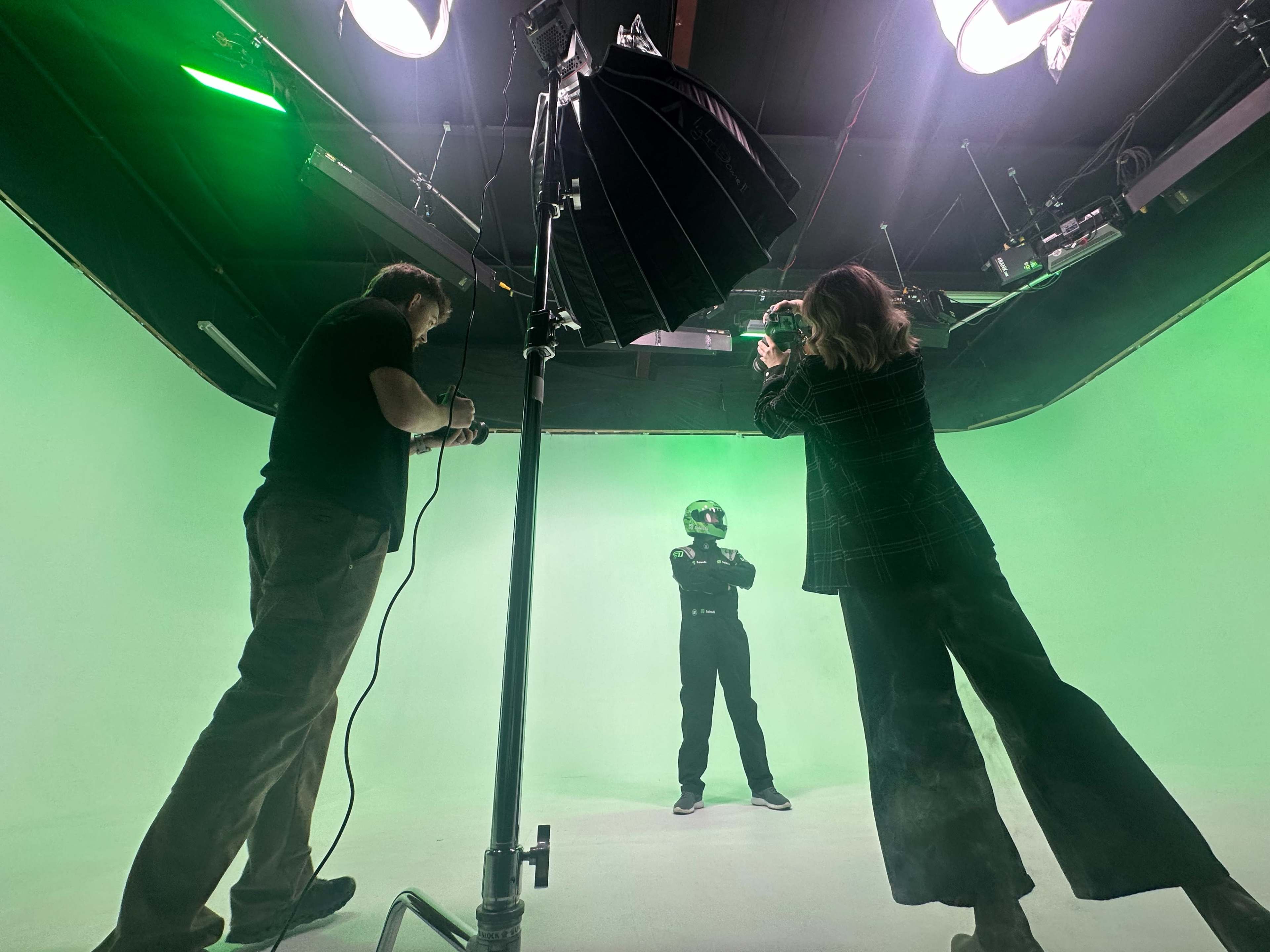 A person in a racing suit and helmet stands in front of a green screen while two crew members adjust lighting and equipment.