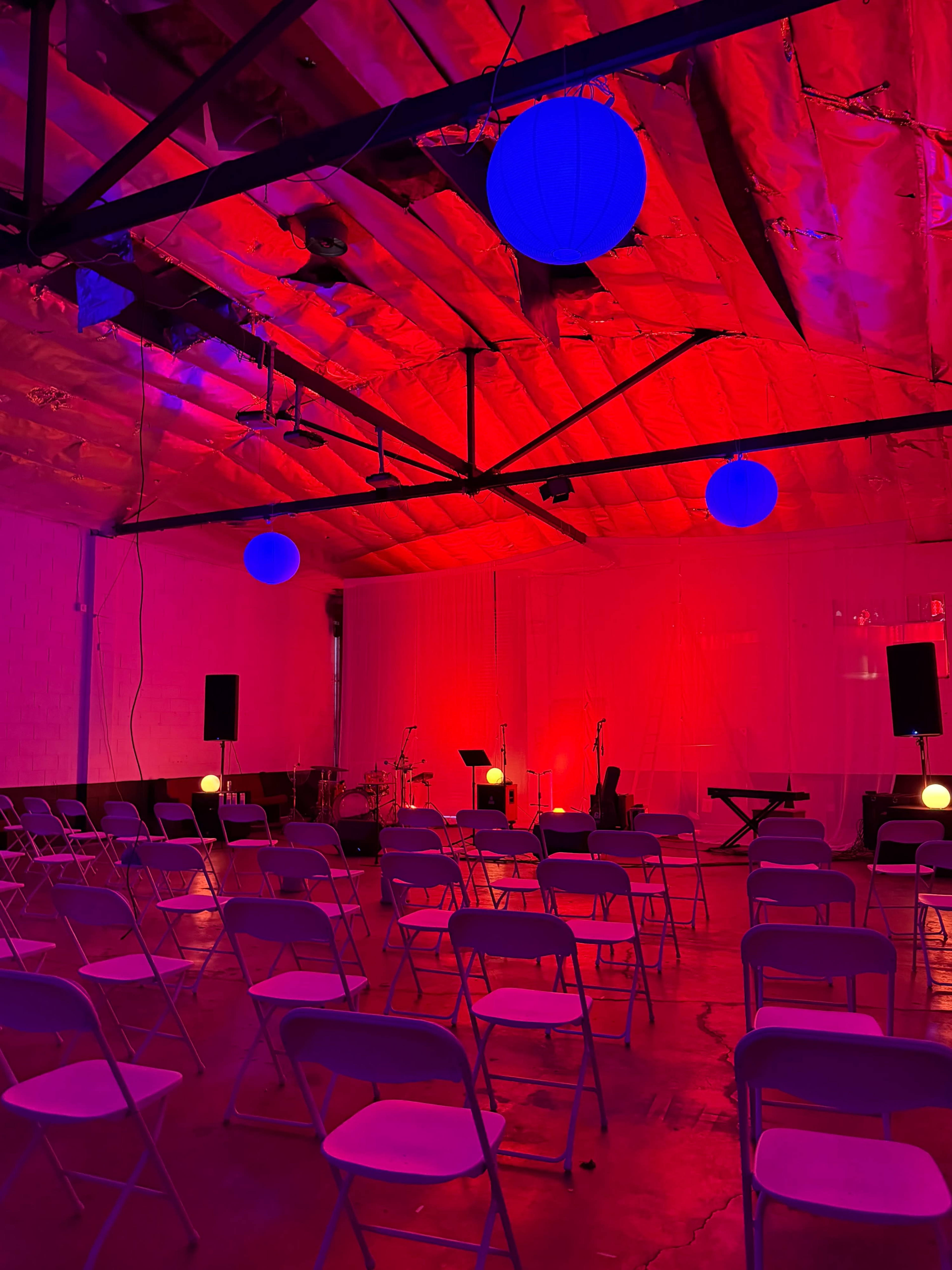 A brightly lit event space features rows of white chairs facing a stage with musical instruments, under red and blue ambient lighting.