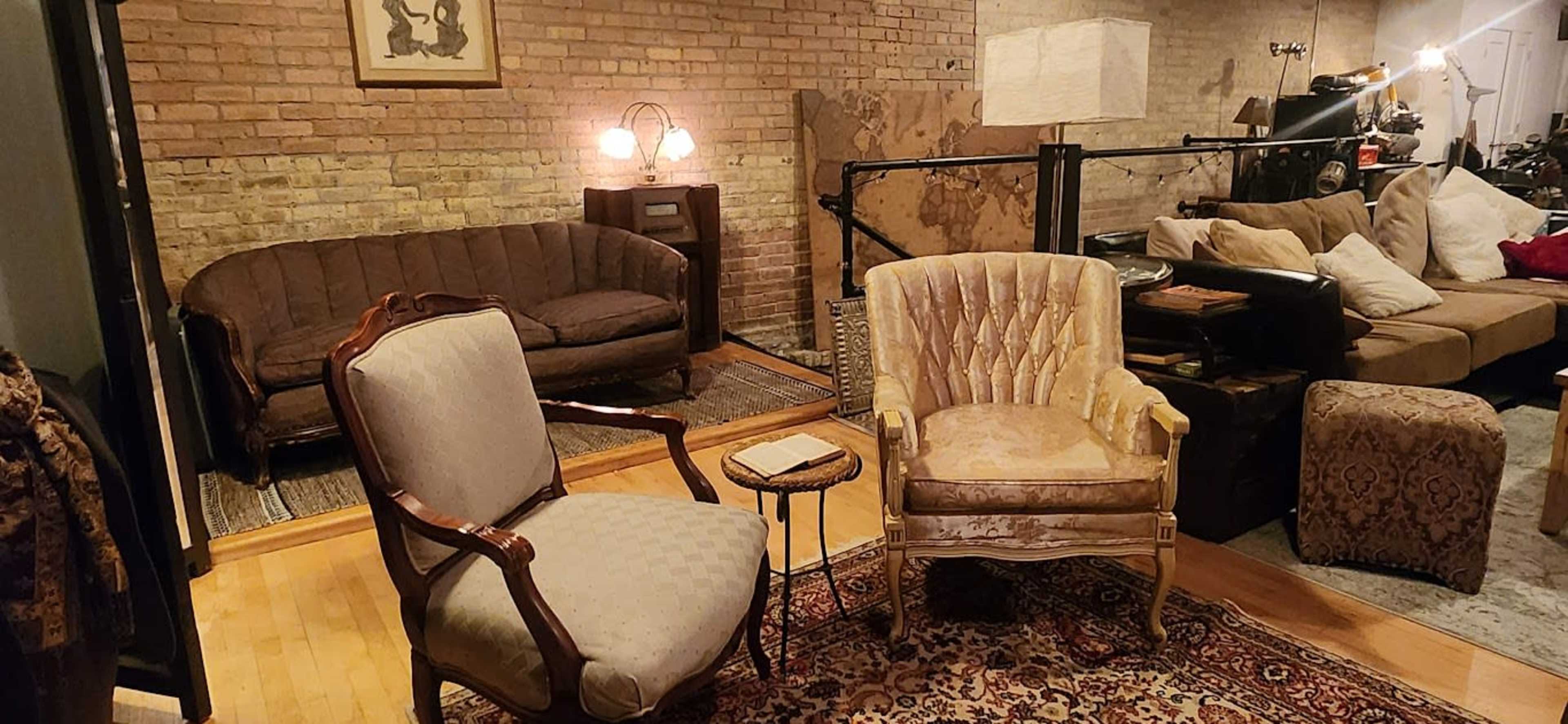 A cozy living room with a brick wall, featuring a vintage armchair and a patterned area rug surrounded by various seating arrangements and decor items.