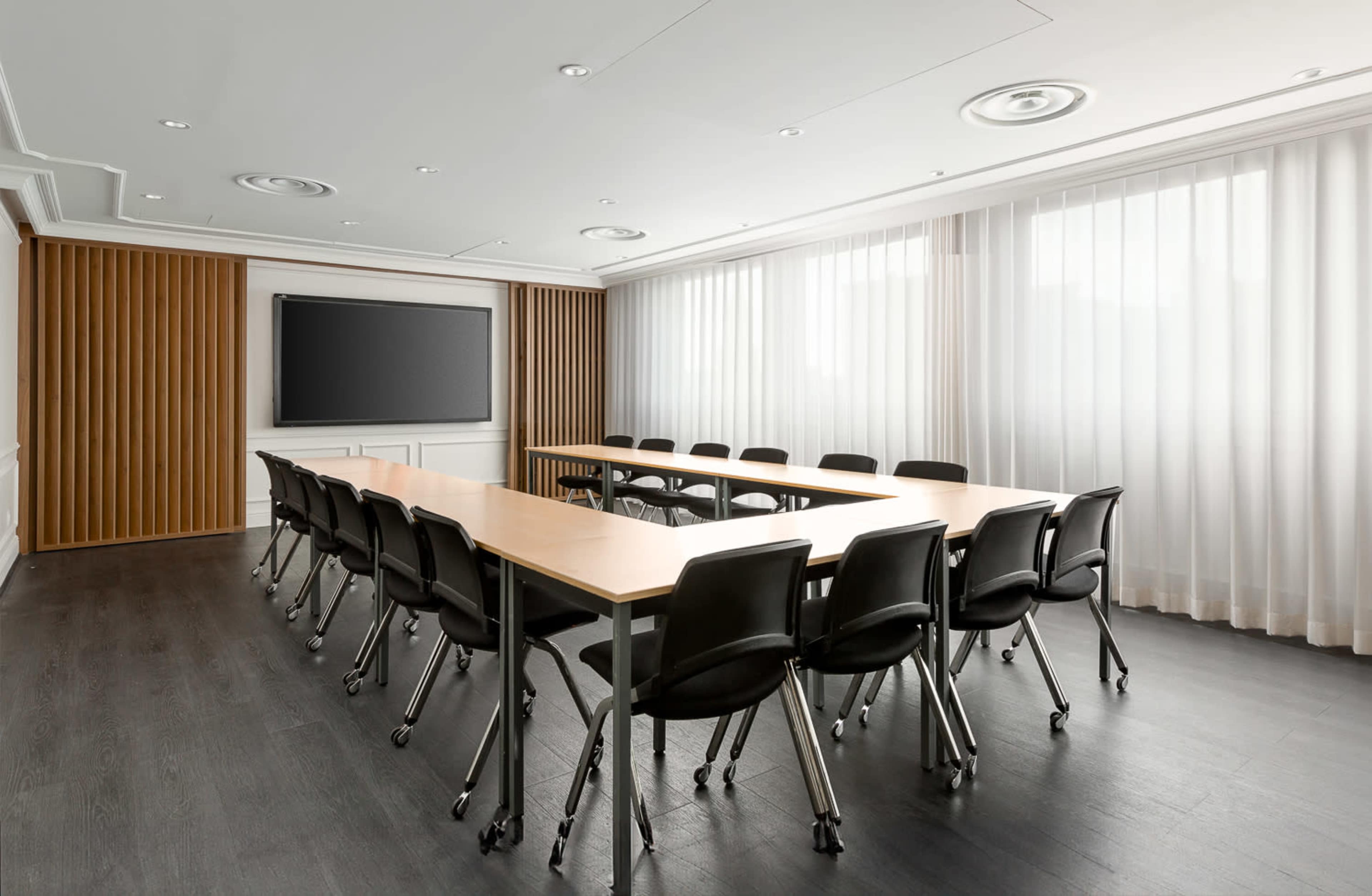 A modern conference room features a long rectangular table surrounded by black chairs and a large screen on the wall, with natural light filtering through sheer curtains.