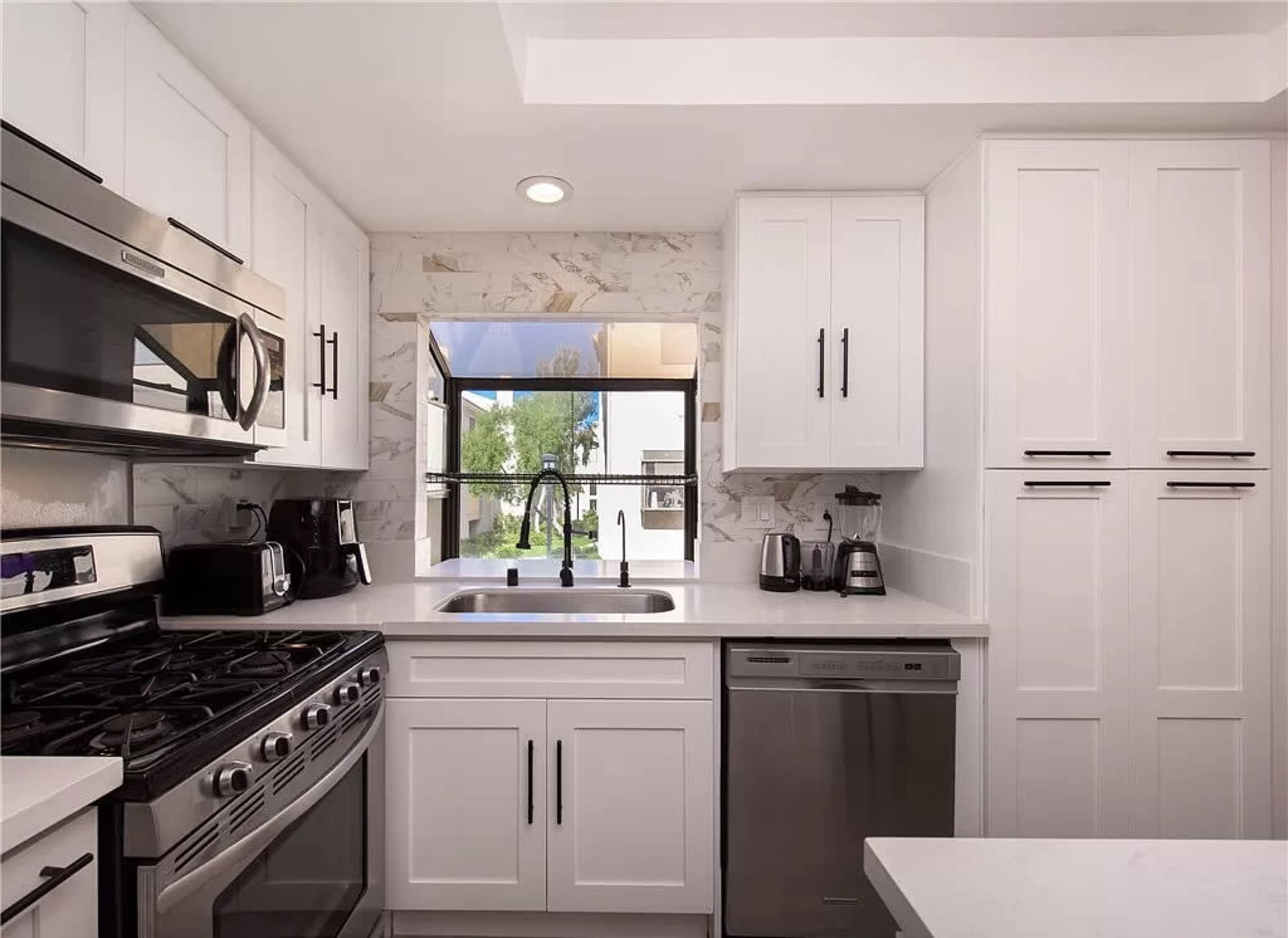 The kitchen features modern white cabinets, stainless steel appliances, and a view of greenery through a large window.