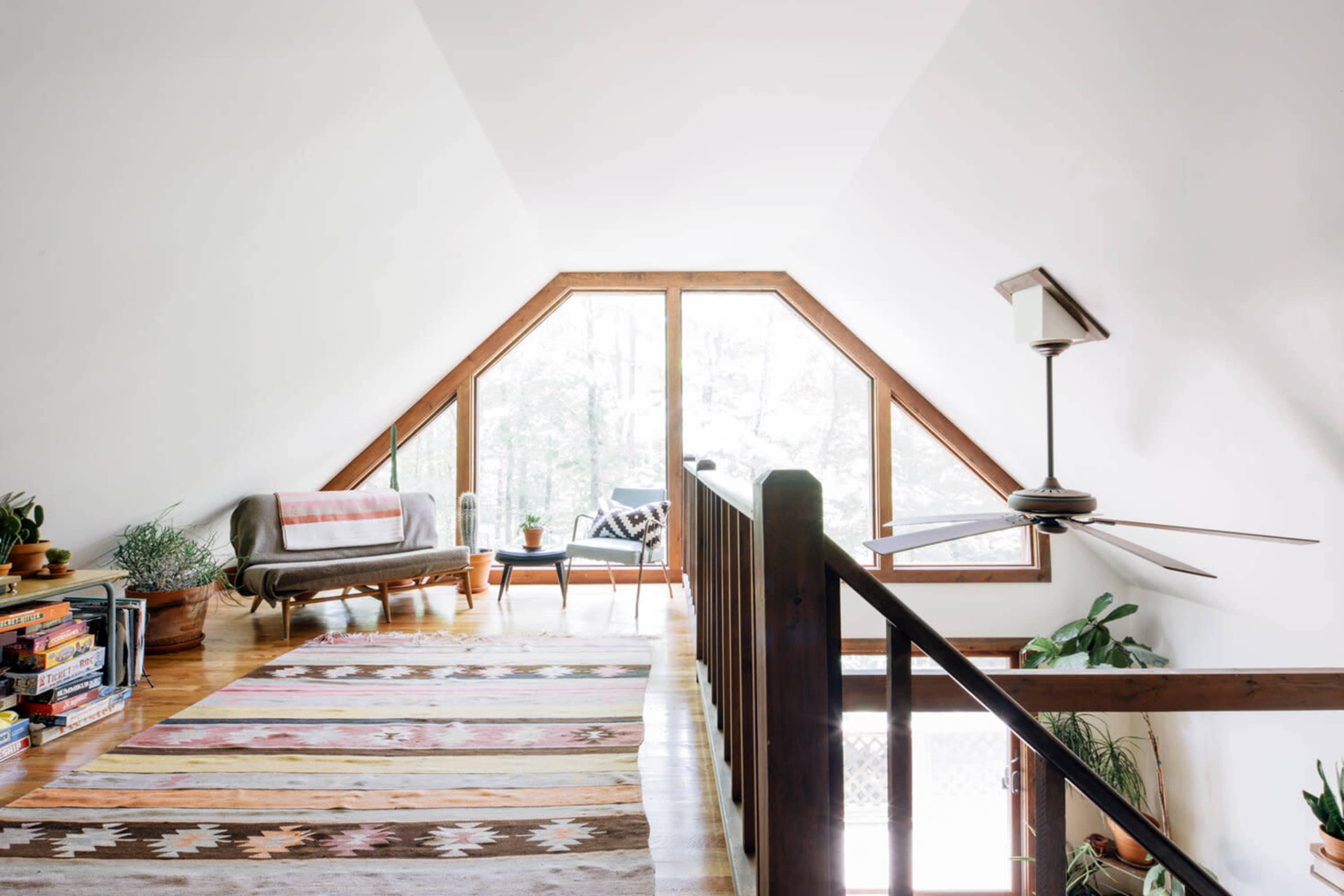 The image shows a bright, triangular attic space with large windows overlooking greenery, furnished with a couch and decorative rugs.