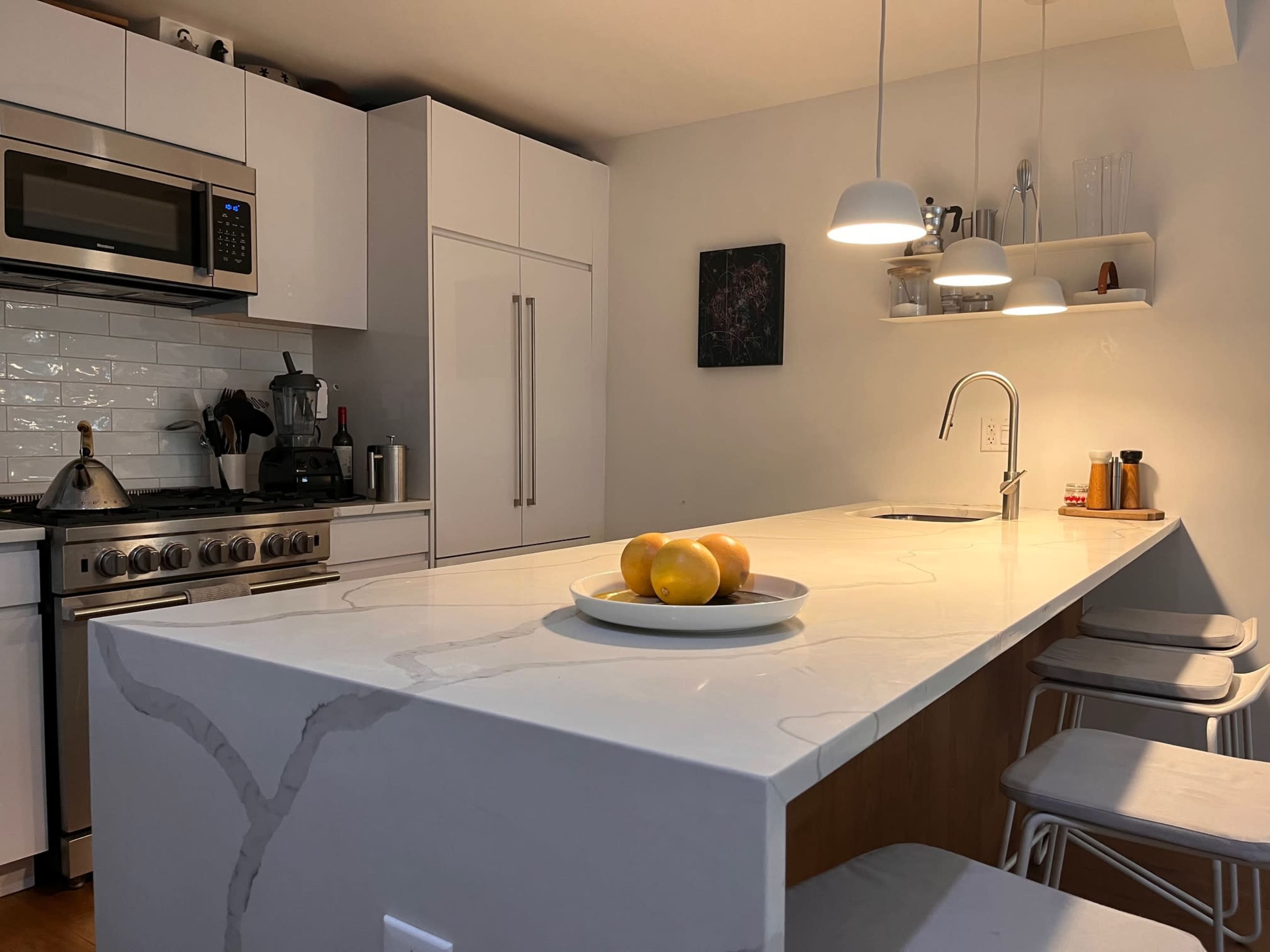 The image shows a modern kitchen with a large island topped with a bowl of oranges, stainless steel appliances, and white cabinetry.