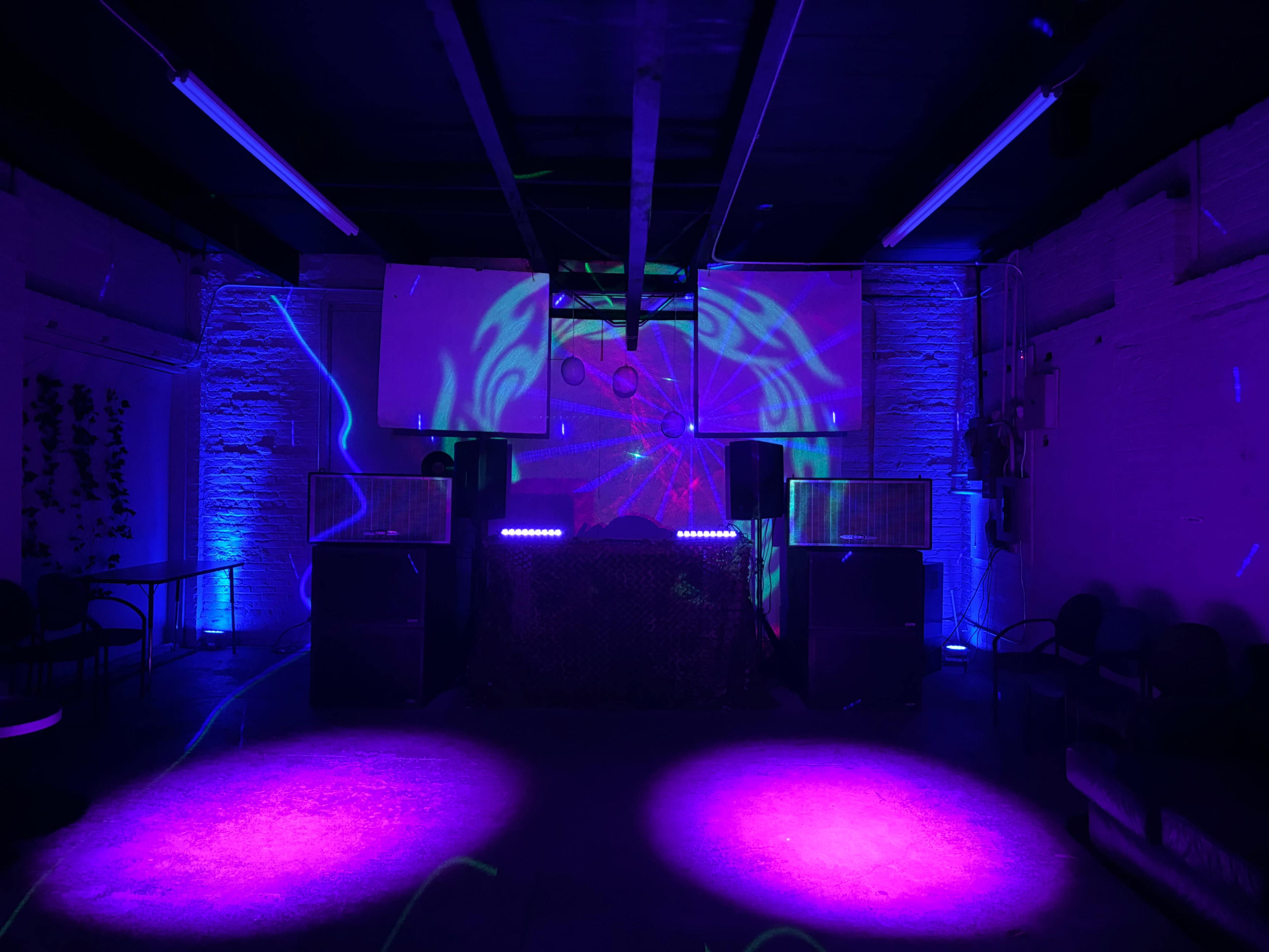 A DJ setup with large speakers and a patterned light display illuminates an empty room with dark walls and seating.