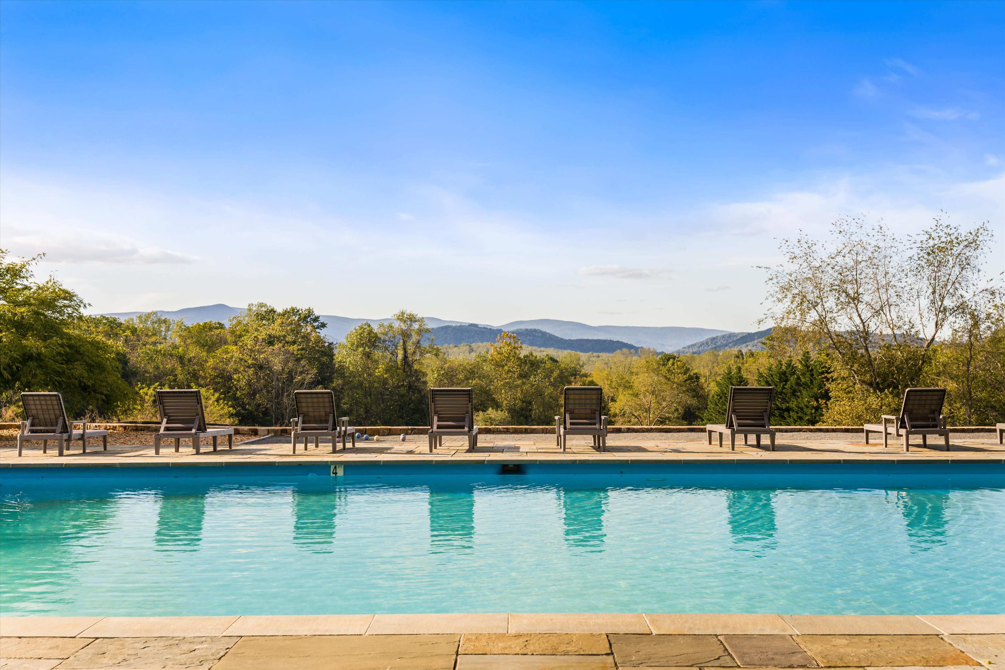 The image shows a swimming pool with several lounge chairs arranged along its edge, set against a backdrop of mountains and trees under a clear blue sky.