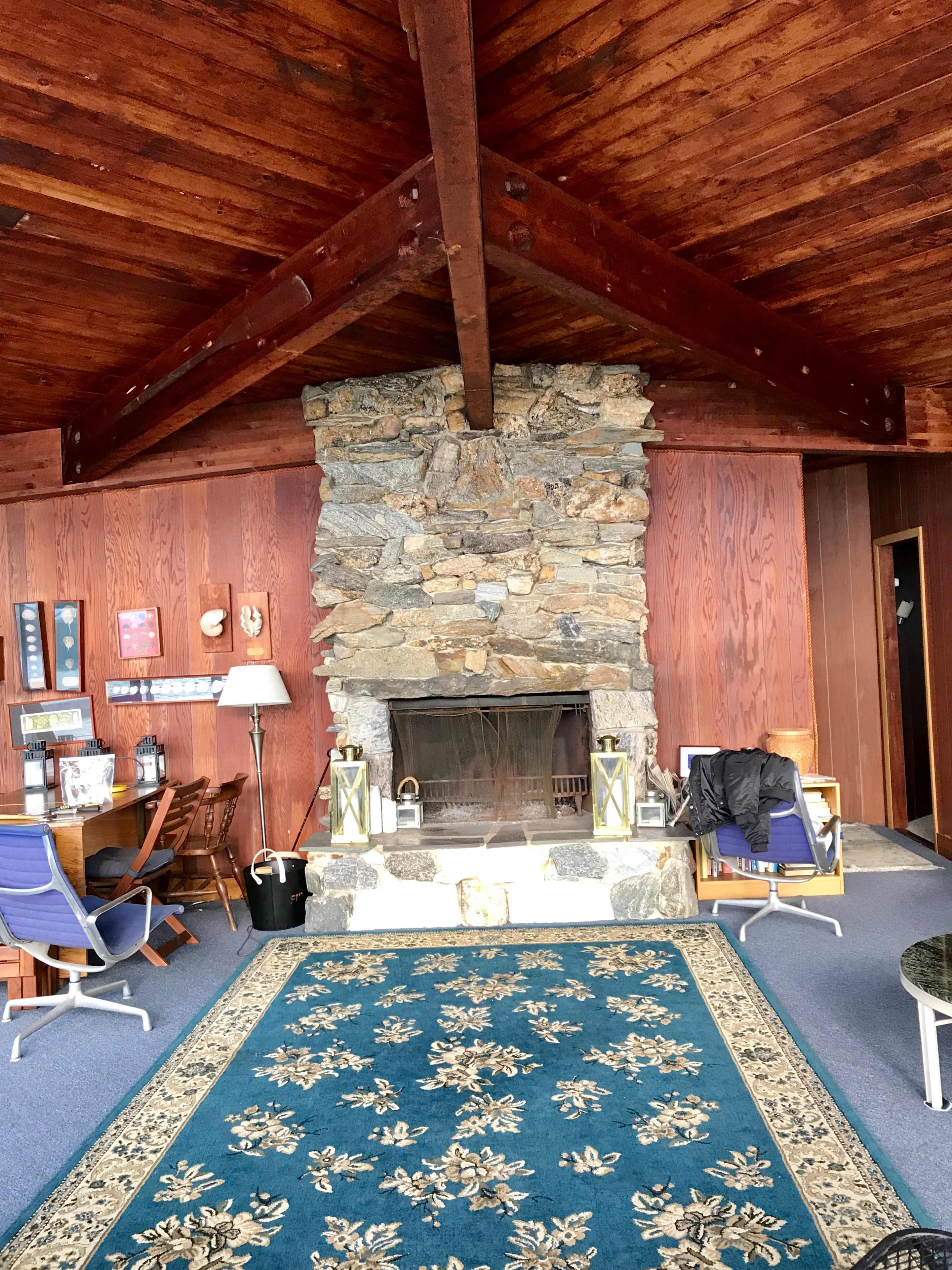 A large stone fireplace is the centerpiece of a room with wooden walls, a wooden ceiling, and a patterned rug on the floor.