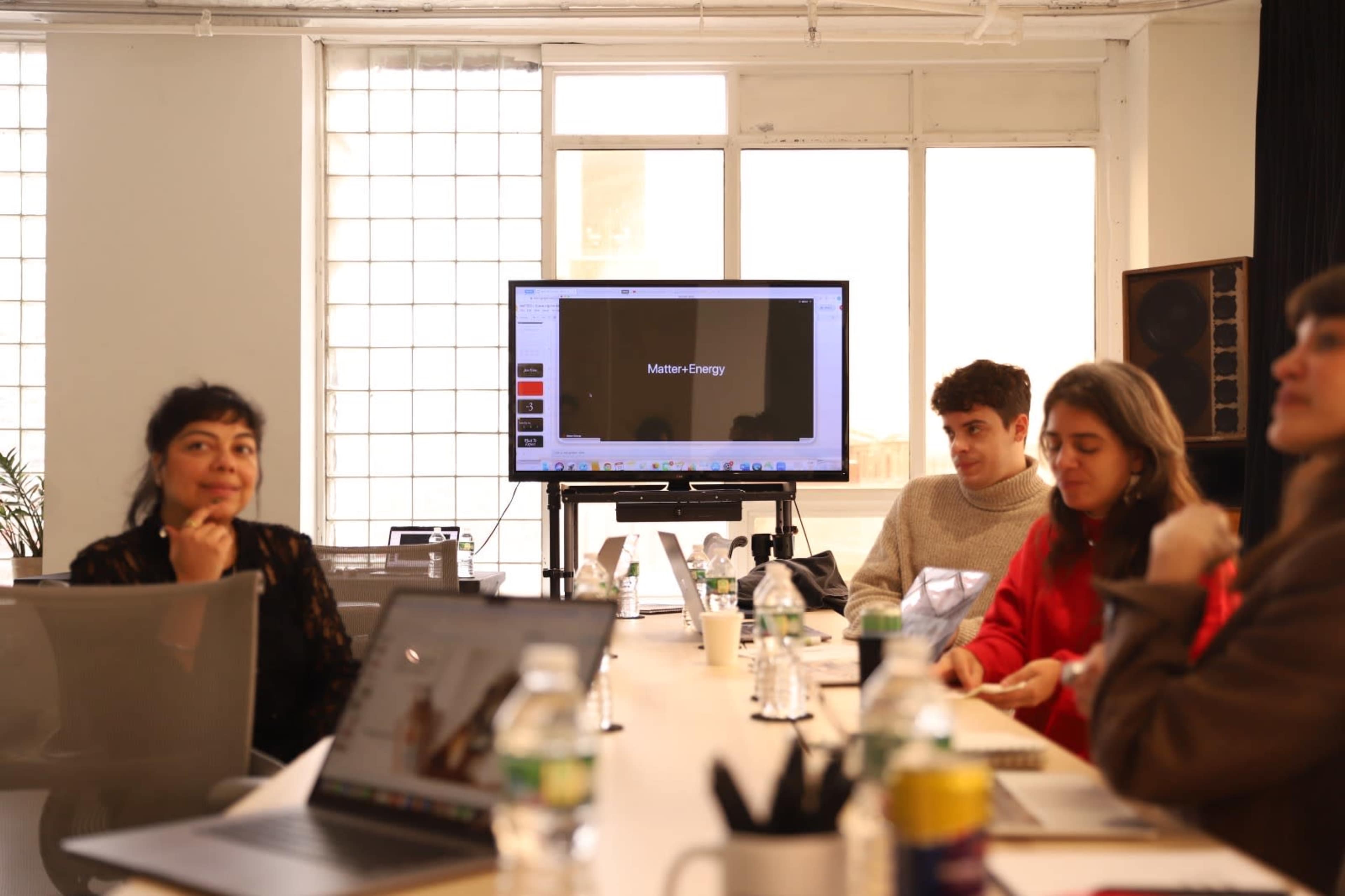 A group of five people is seated around a table during a meeting, with a presentation screen displaying a title in the background.