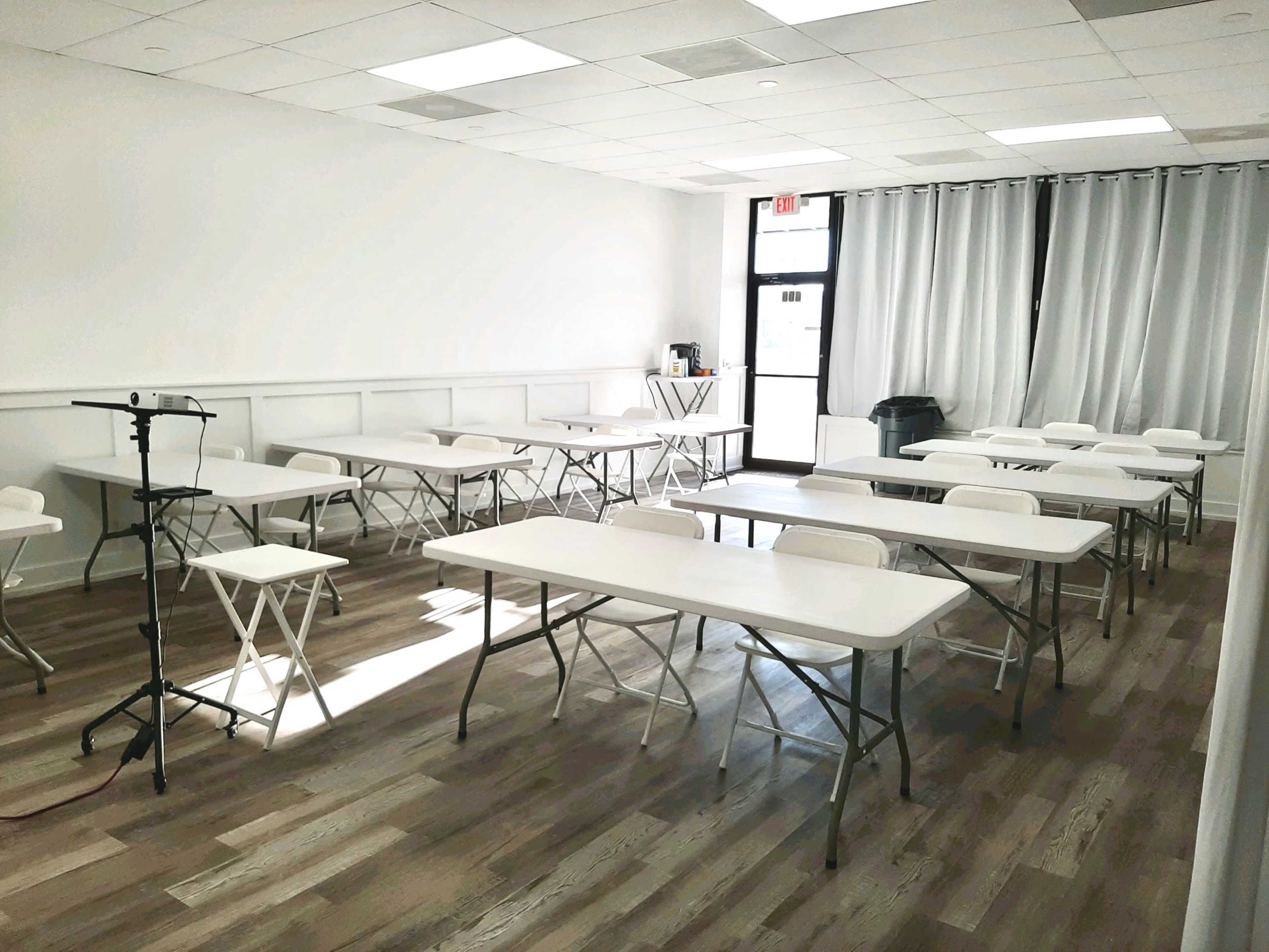 A bright, empty classroom with several white folding tables and chairs arranged in rows, and a microphone stand positioned in one corner.