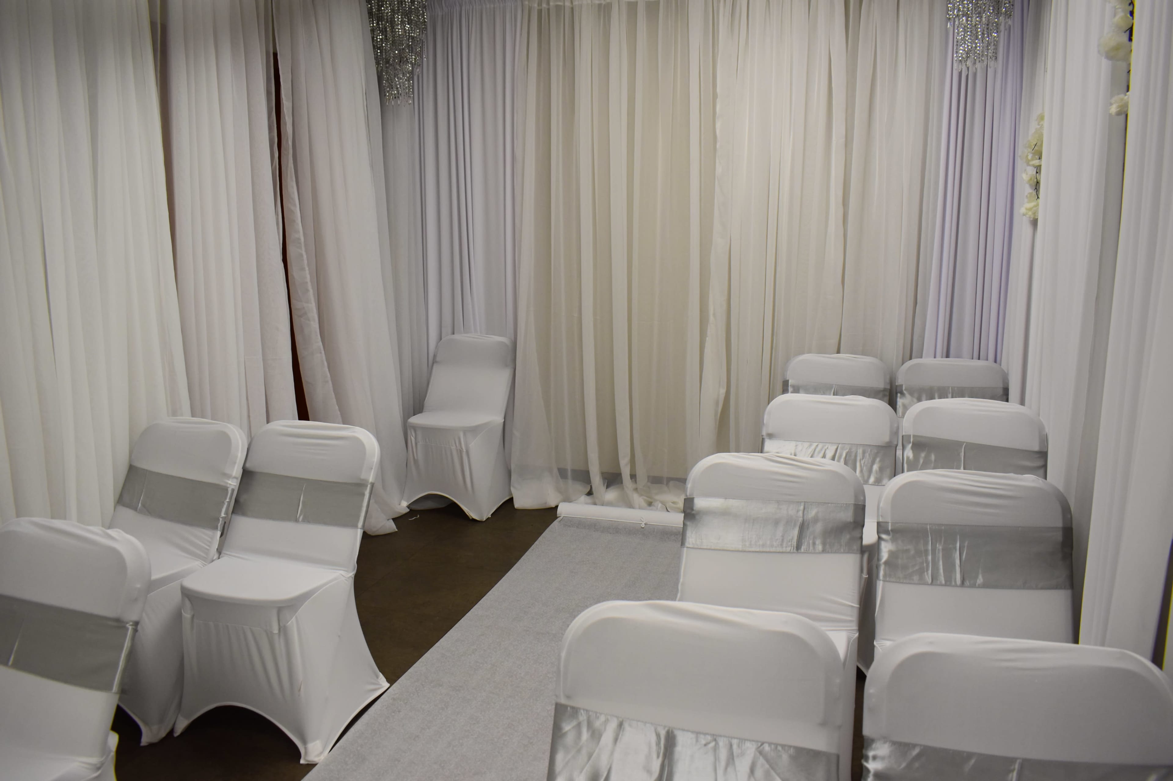 The room features a row of white chairs with silver sashes arranged on either side of a carpeted aisle, surrounded by white draped curtains.