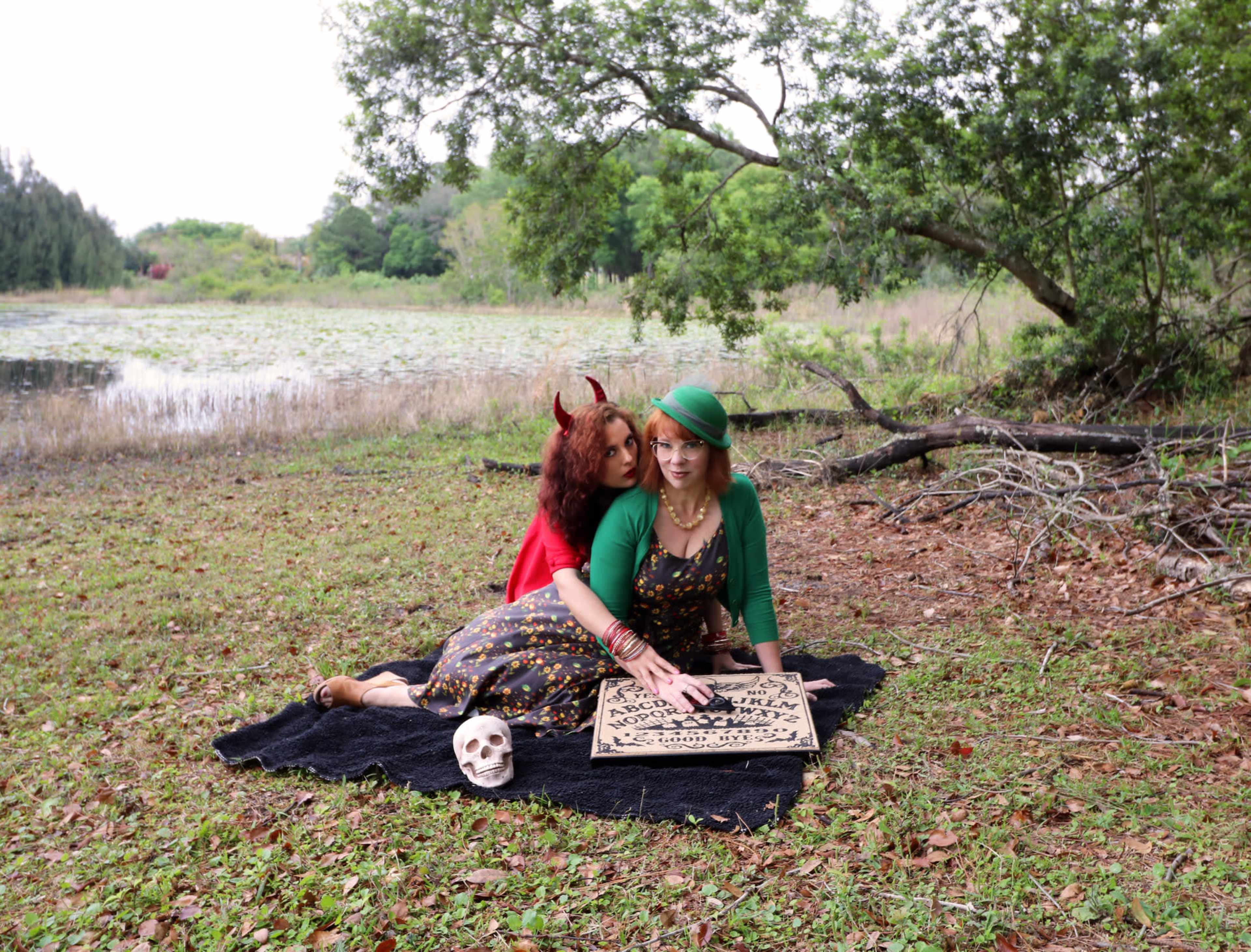 Two women are seated on a blanket in a natural setting by a body of water, one dressed in a devil costume and the other in a green outfit, as they engage with a Ouija board.