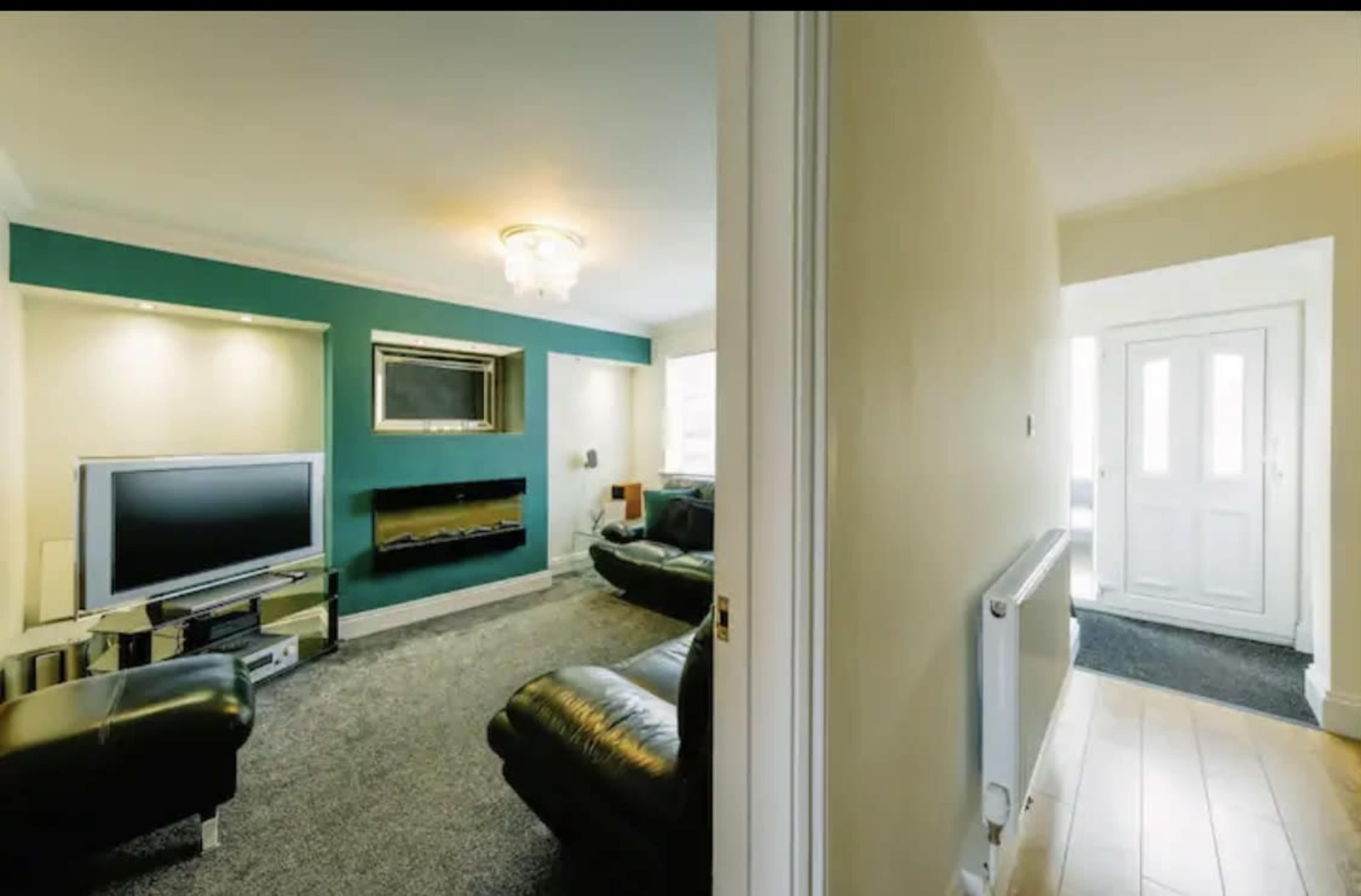 A modern living room with a television and a fireplace, adjacent to a hallway that leads to the front door.