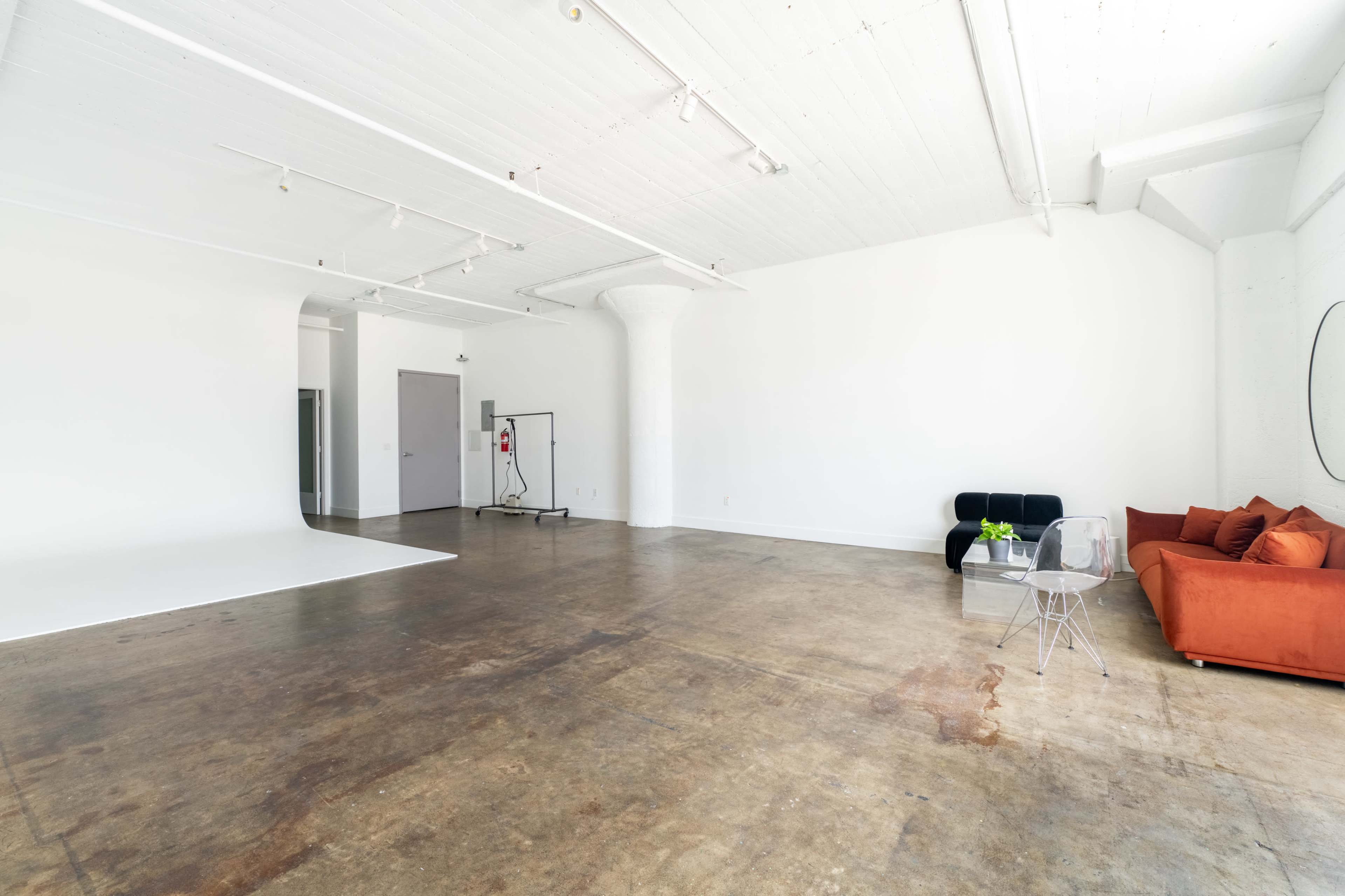 The image shows a spacious, minimalist studio with a concrete floor, a white backdrop, and a modern orange couch accompanied by a clear acrylic chair.