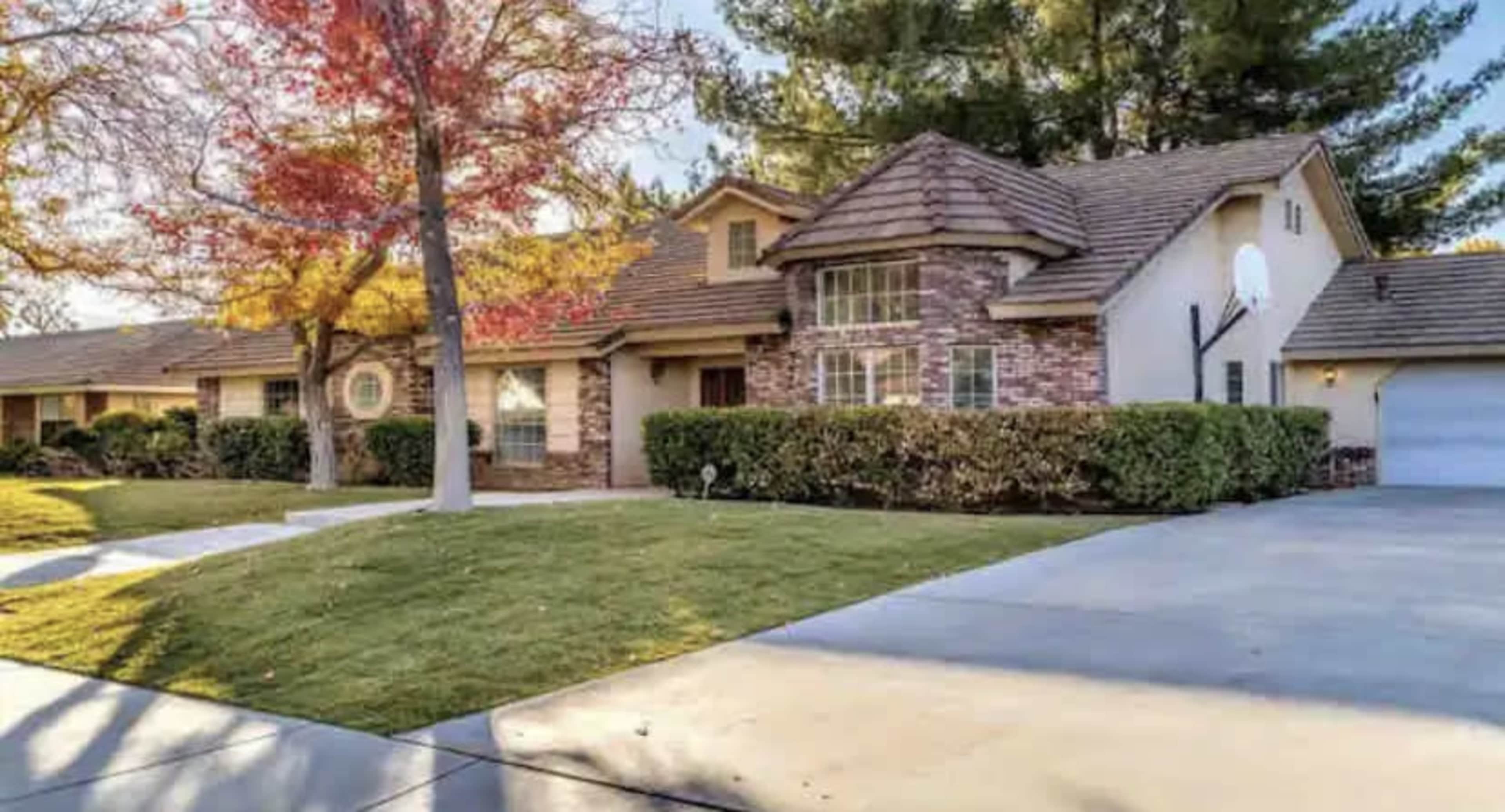 A single-story brick house with a sloped roof, surrounded by well-maintained lawns and trees, with a driveway leading to a garage.