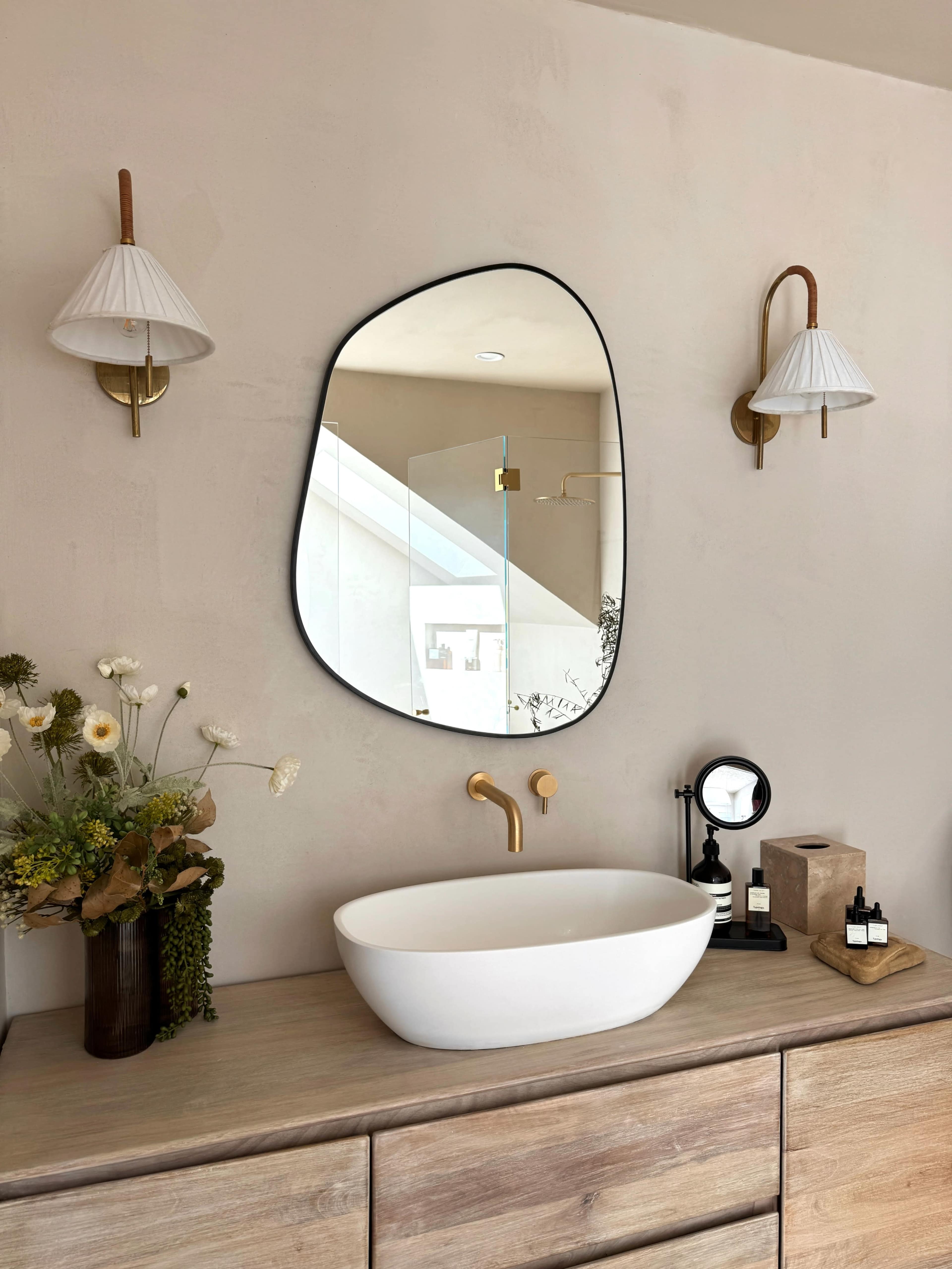 A modern bathroom vanity with a large, irregularly-shaped mirror, a white vessel sink, brass fixtures, and decorative items, including a flower arrangement and various bottles.