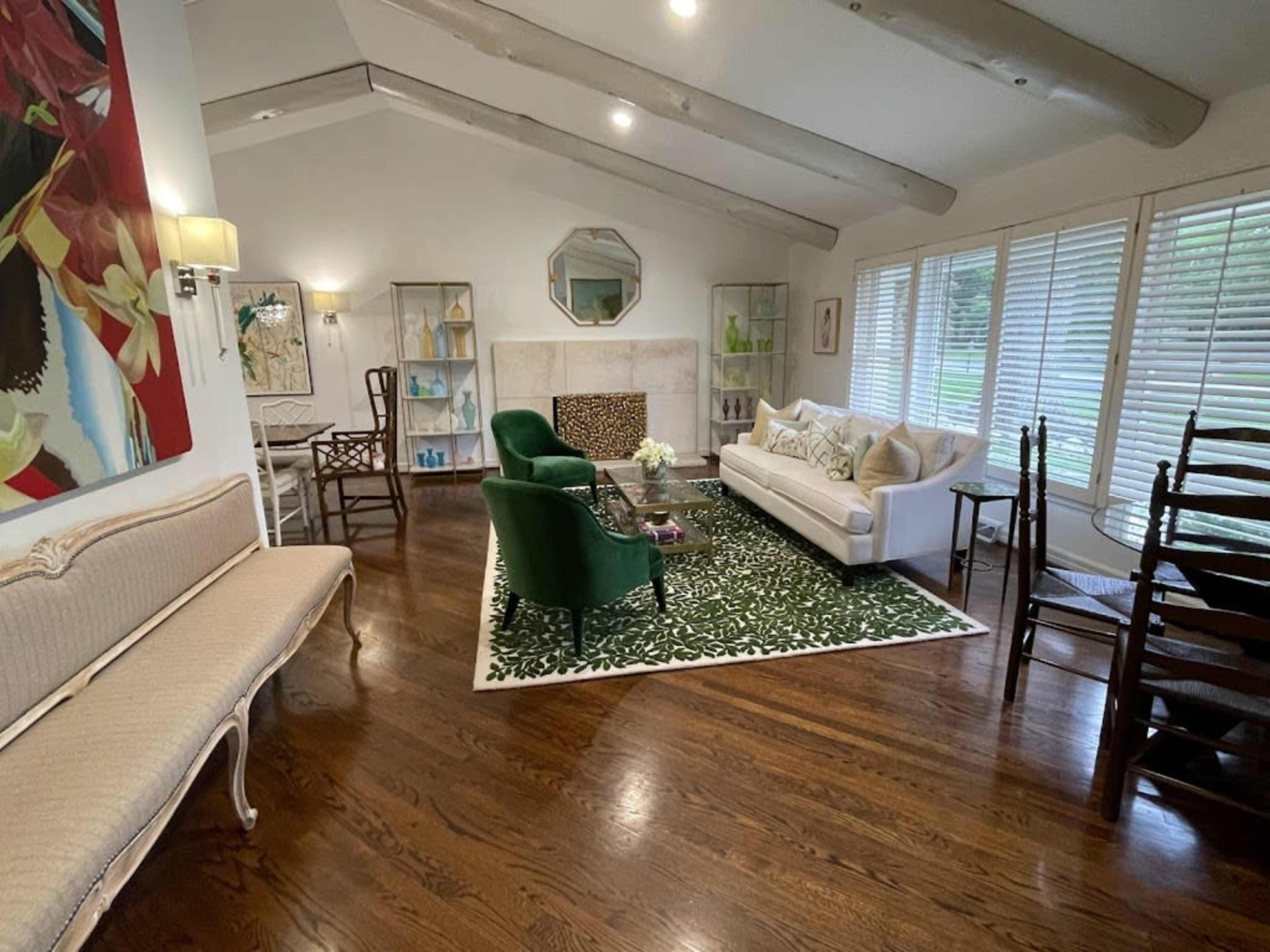 A modern living room features a white sofa, green armchairs, and a decorative rug, with large windows and wooden beams adding to the airy atmosphere.