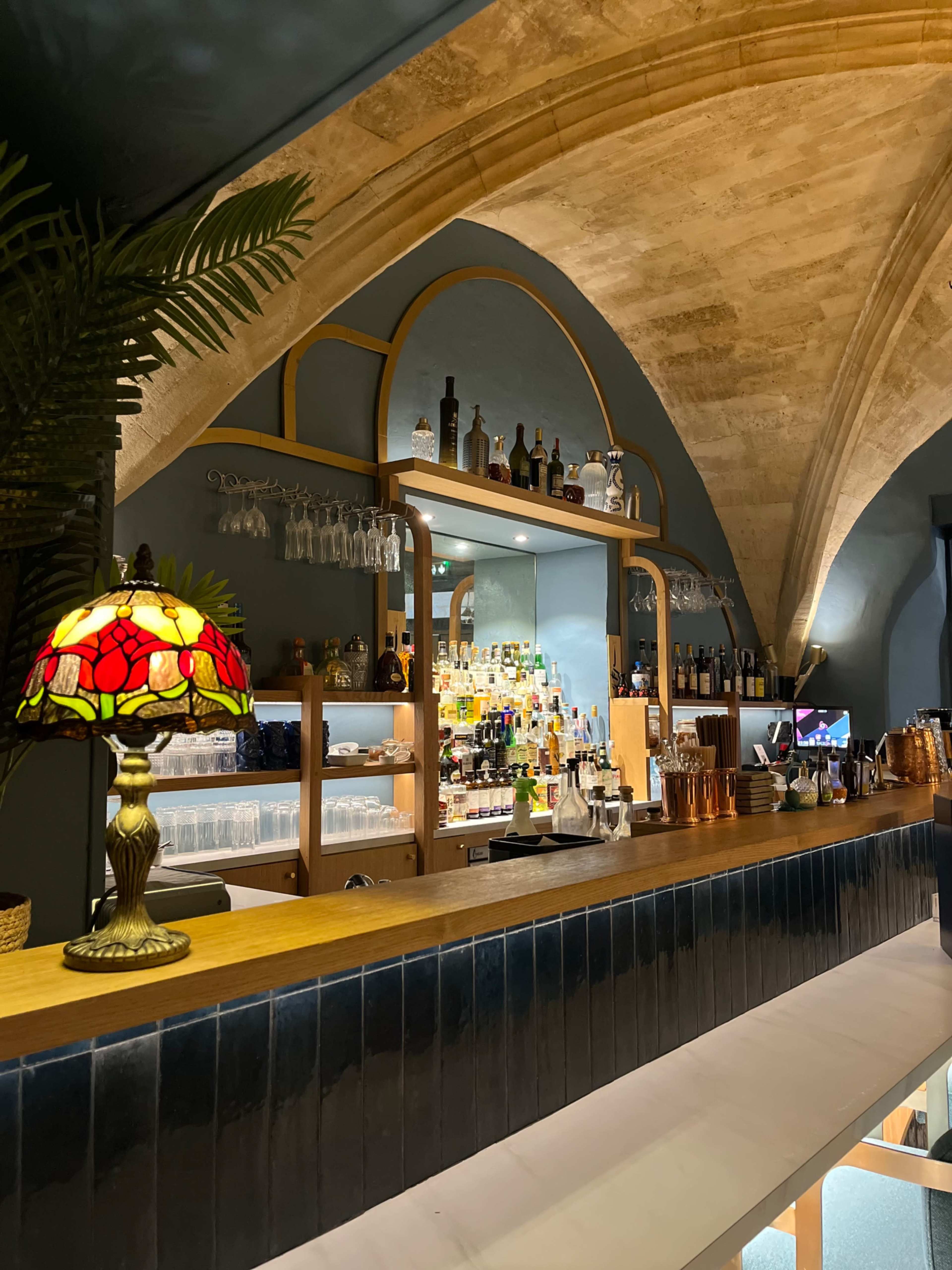 The image shows a bar with an arched ceiling, shelves filled with bottles, and a decorative lamp on the counter.