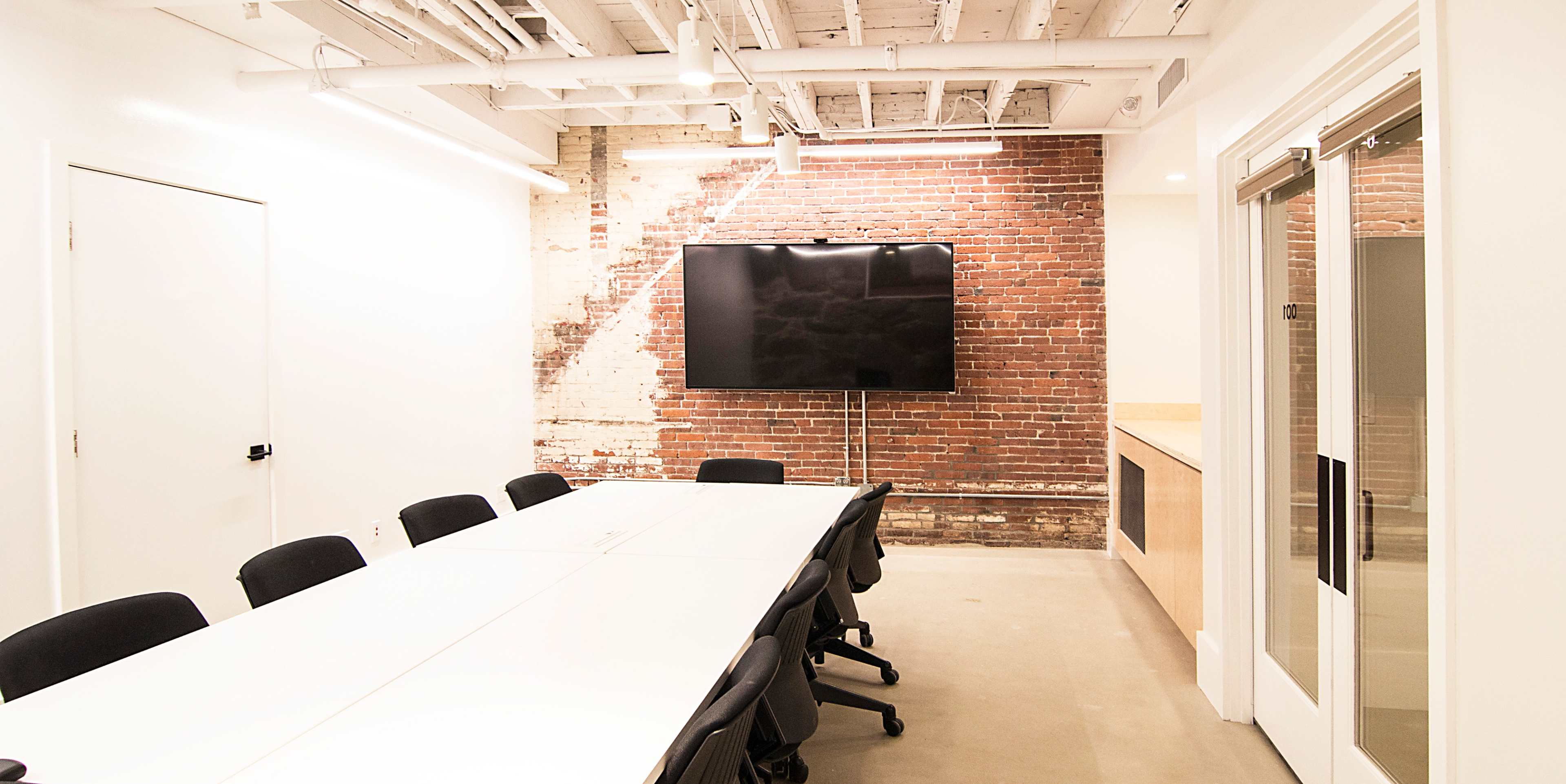 A conference room features a long white table surrounded by black chairs and a large flat-screen TV mounted on a brick wall.
