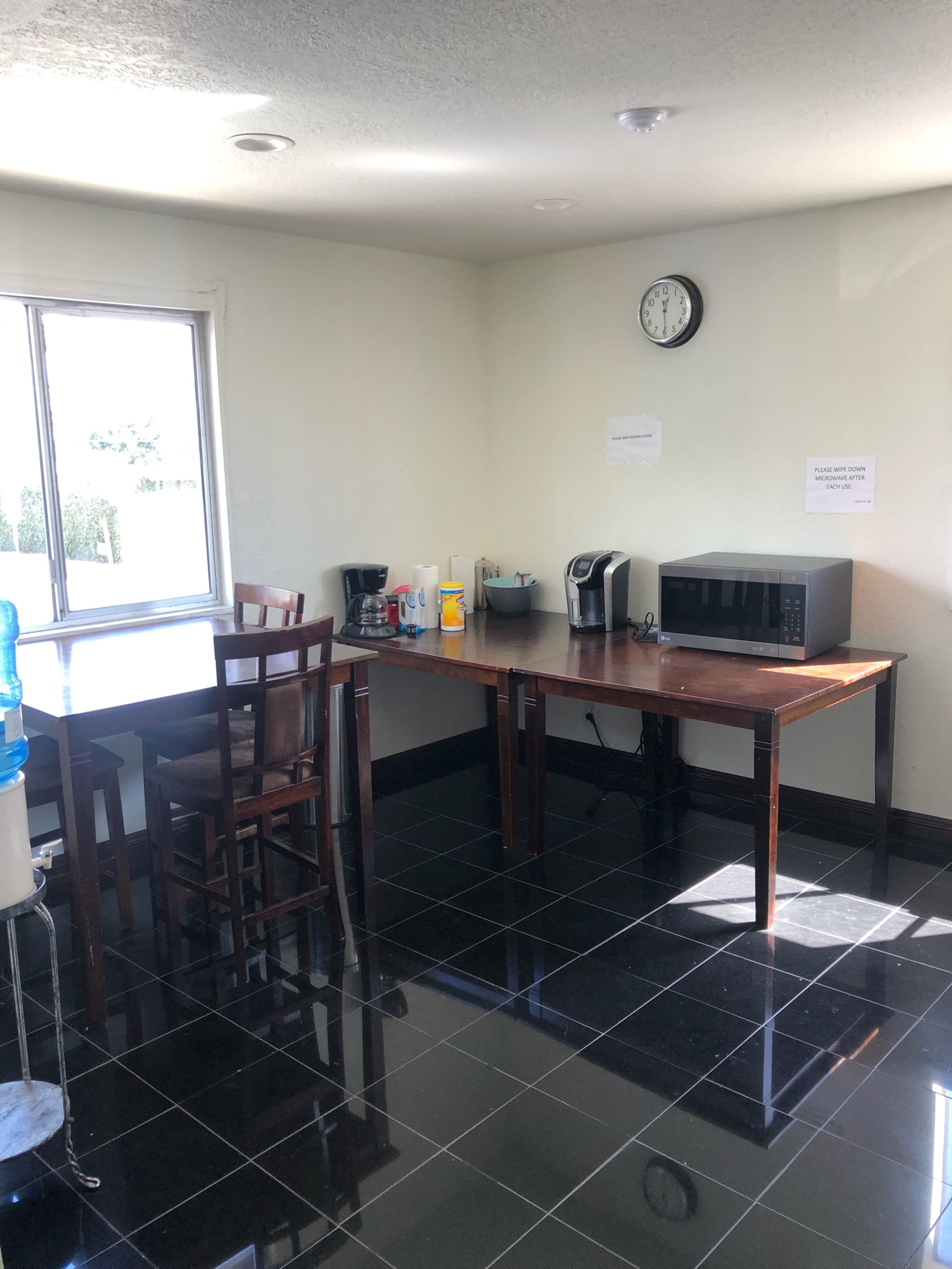 A simple kitchen area with two tables, a water cooler, a coffee maker, a microwave, and a clock on the wall.