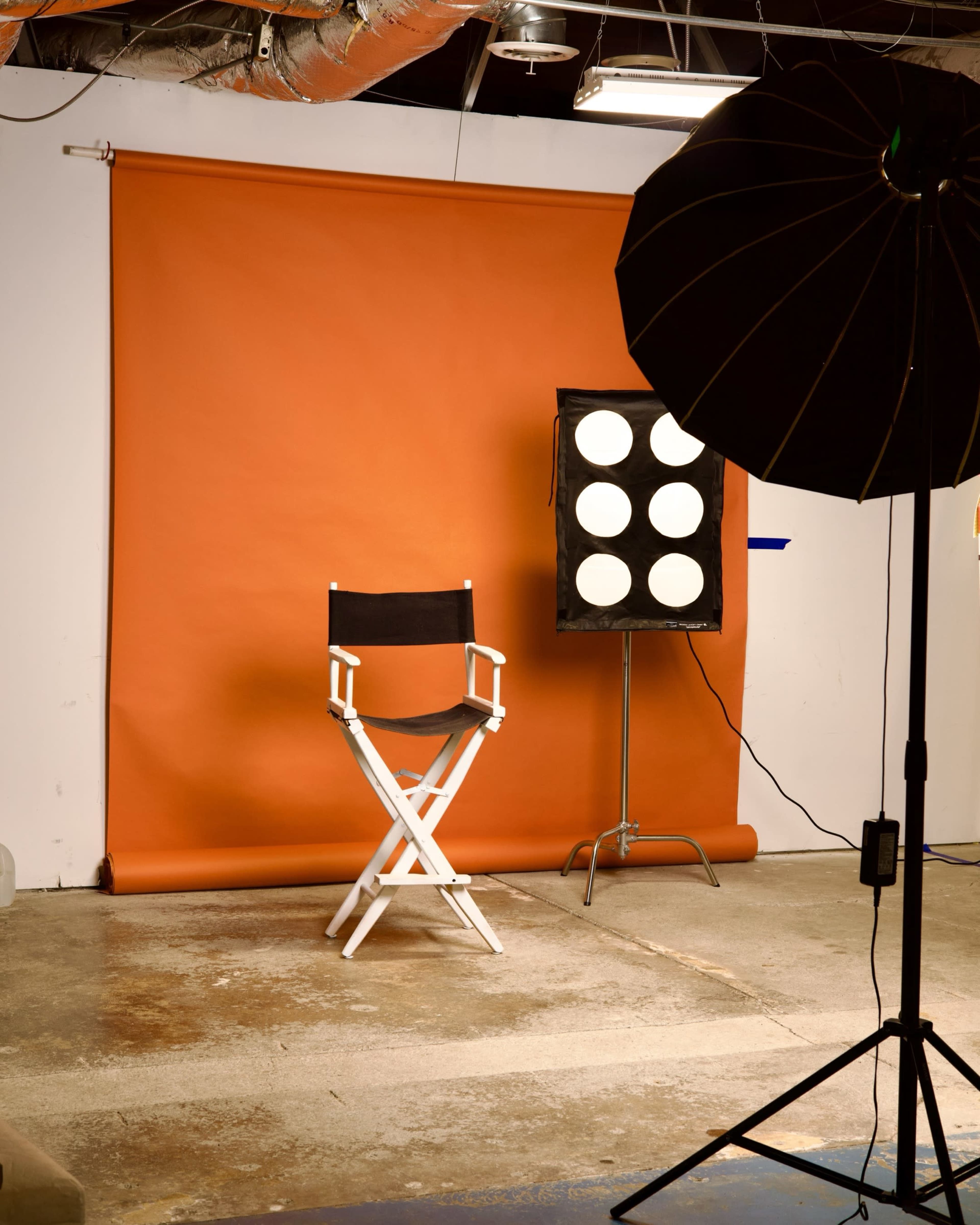 A photography studio features an orange backdrop, a director's chair, and a light setup with multiple circular lamps.
