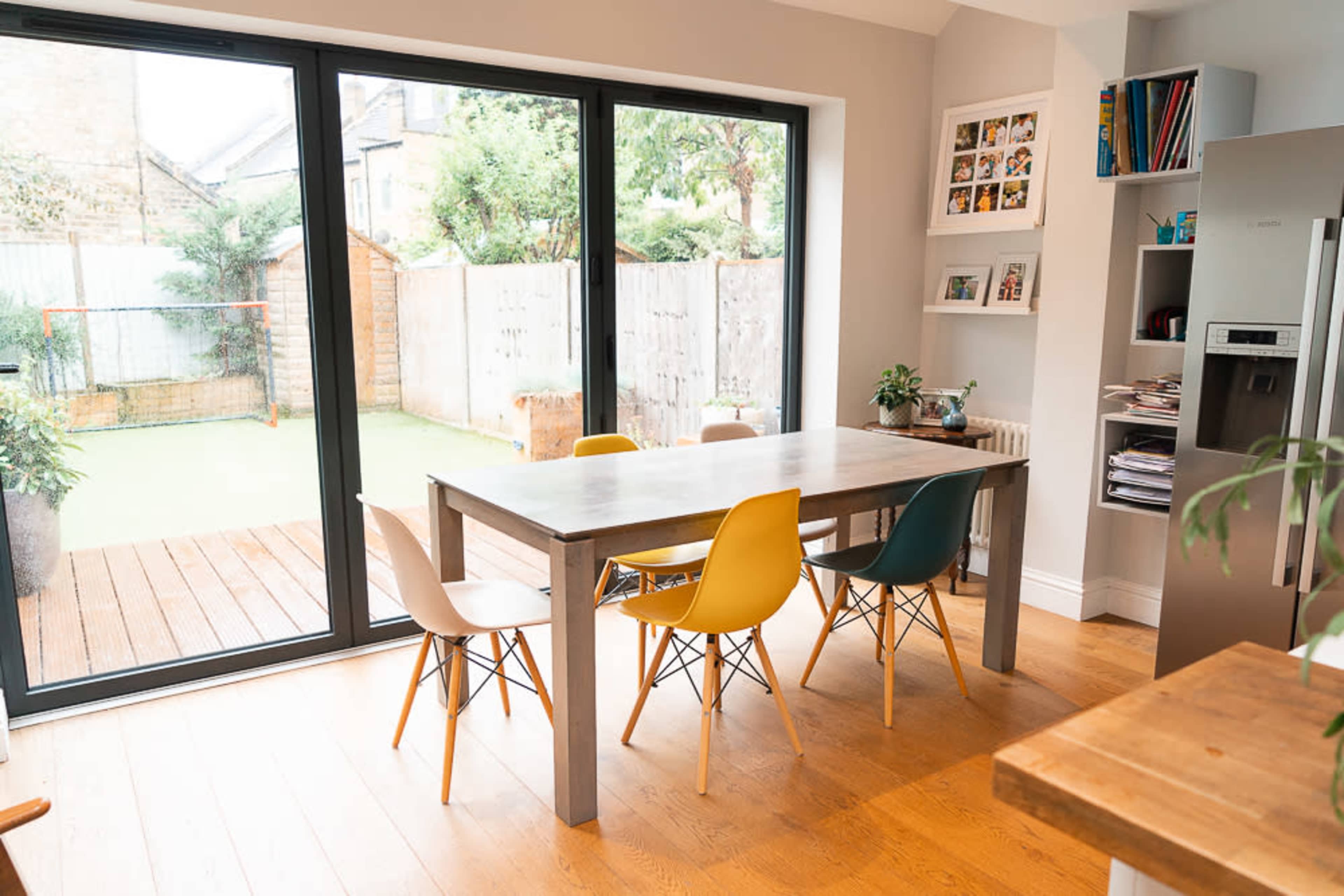 A dining area features a wooden table surrounded by colorful chairs, with large glass doors opening to a backyard.