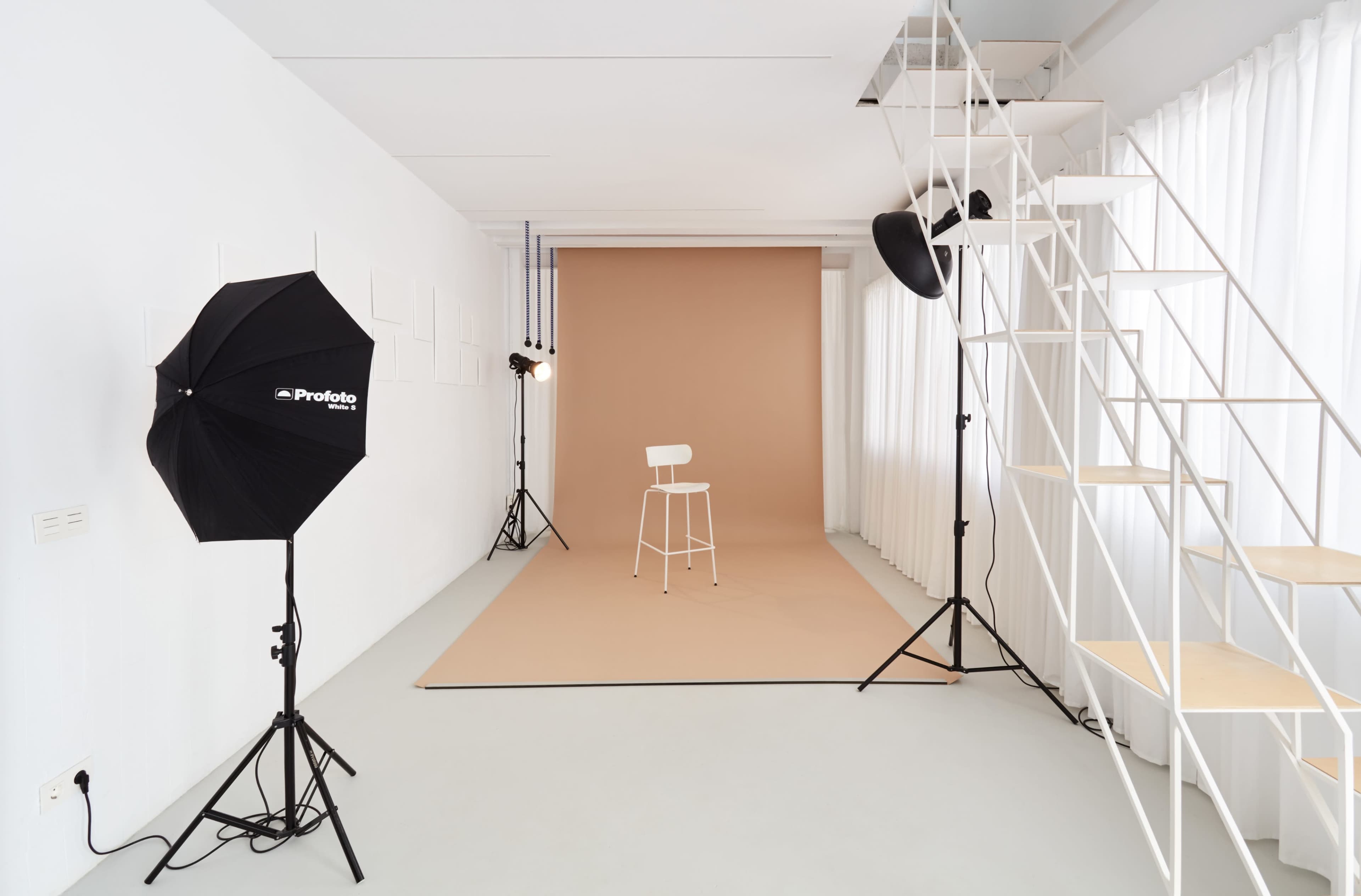 The image shows a photography studio with a beige backdrop, a white stool in the center, and studio lights positioned on either side.