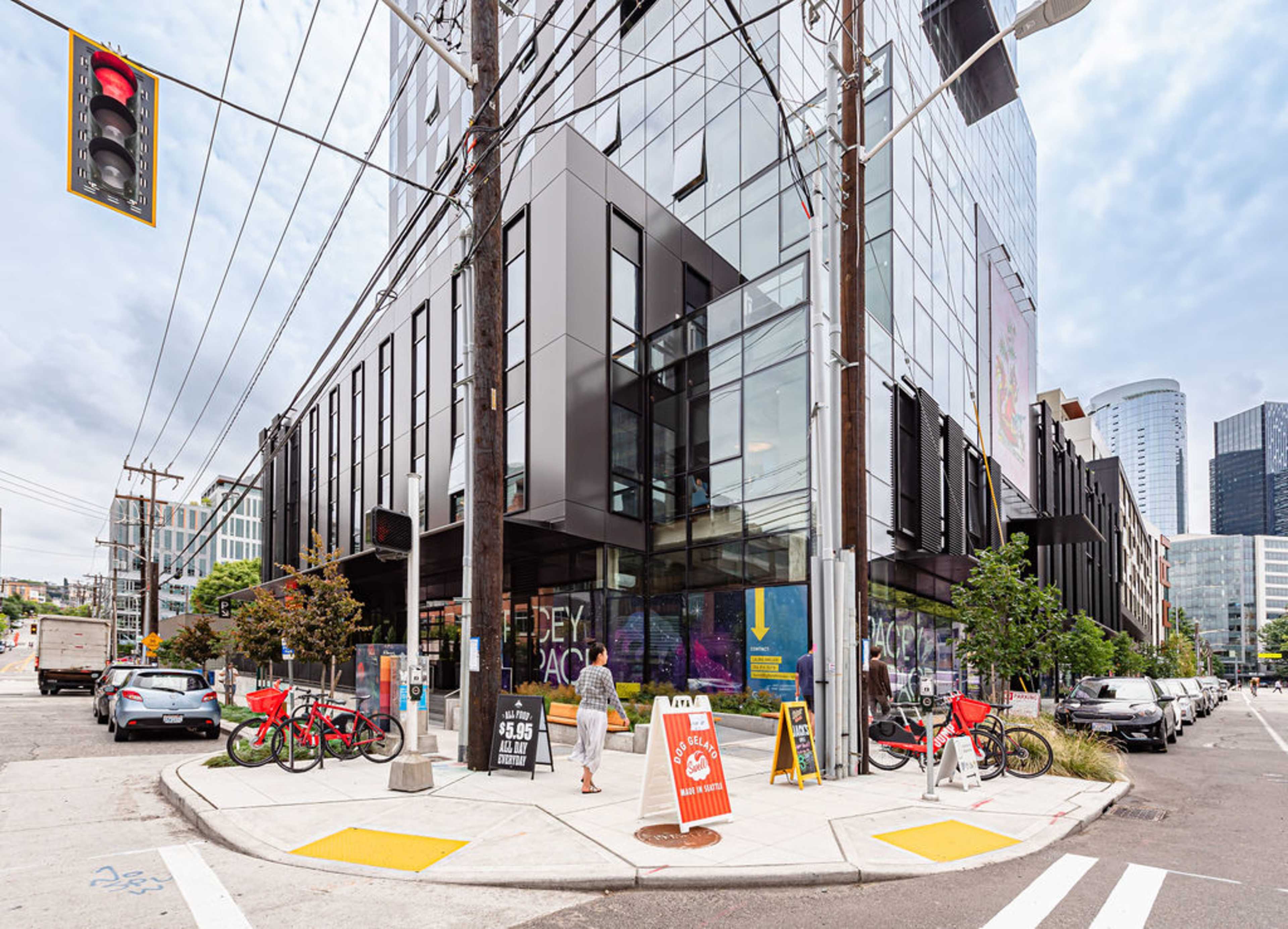 A modern building with large glass windows and a corner entrance is situated at a busy intersection, surrounded by parked cars and bicycles.
