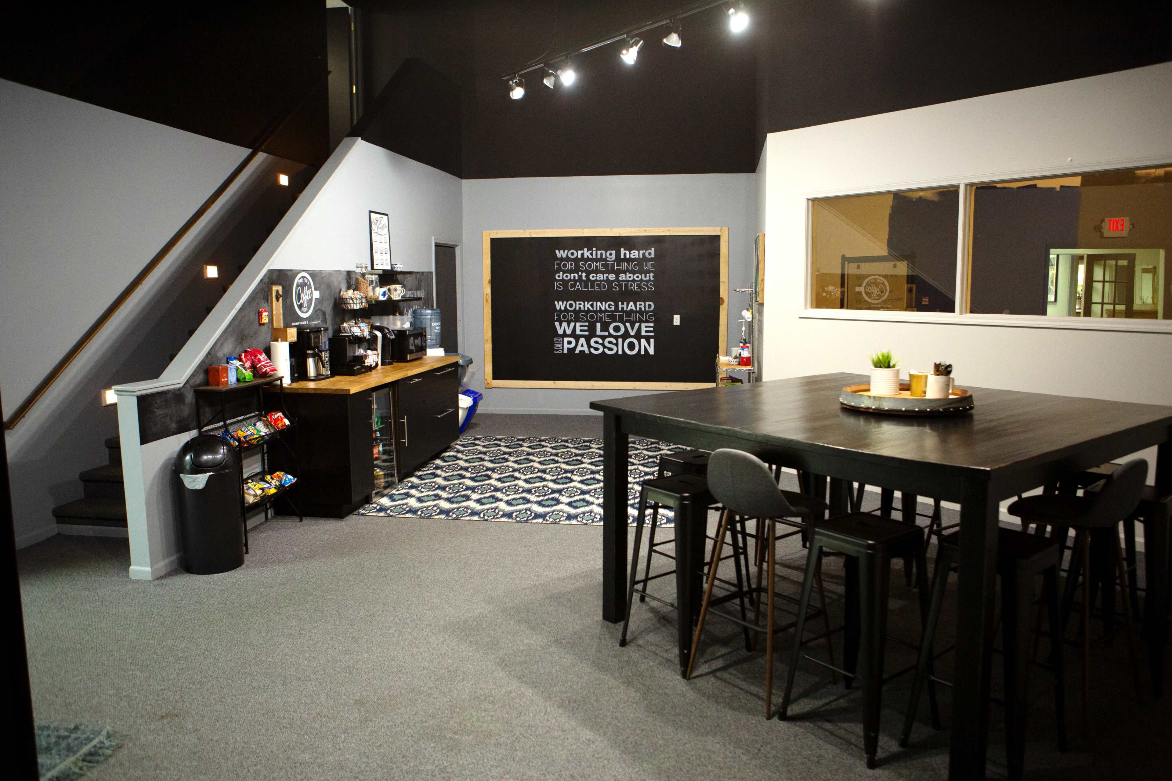 A modern office break room featuring a coffee station, snack area, and a large table with high chairs, accented by a motivational wall mural.