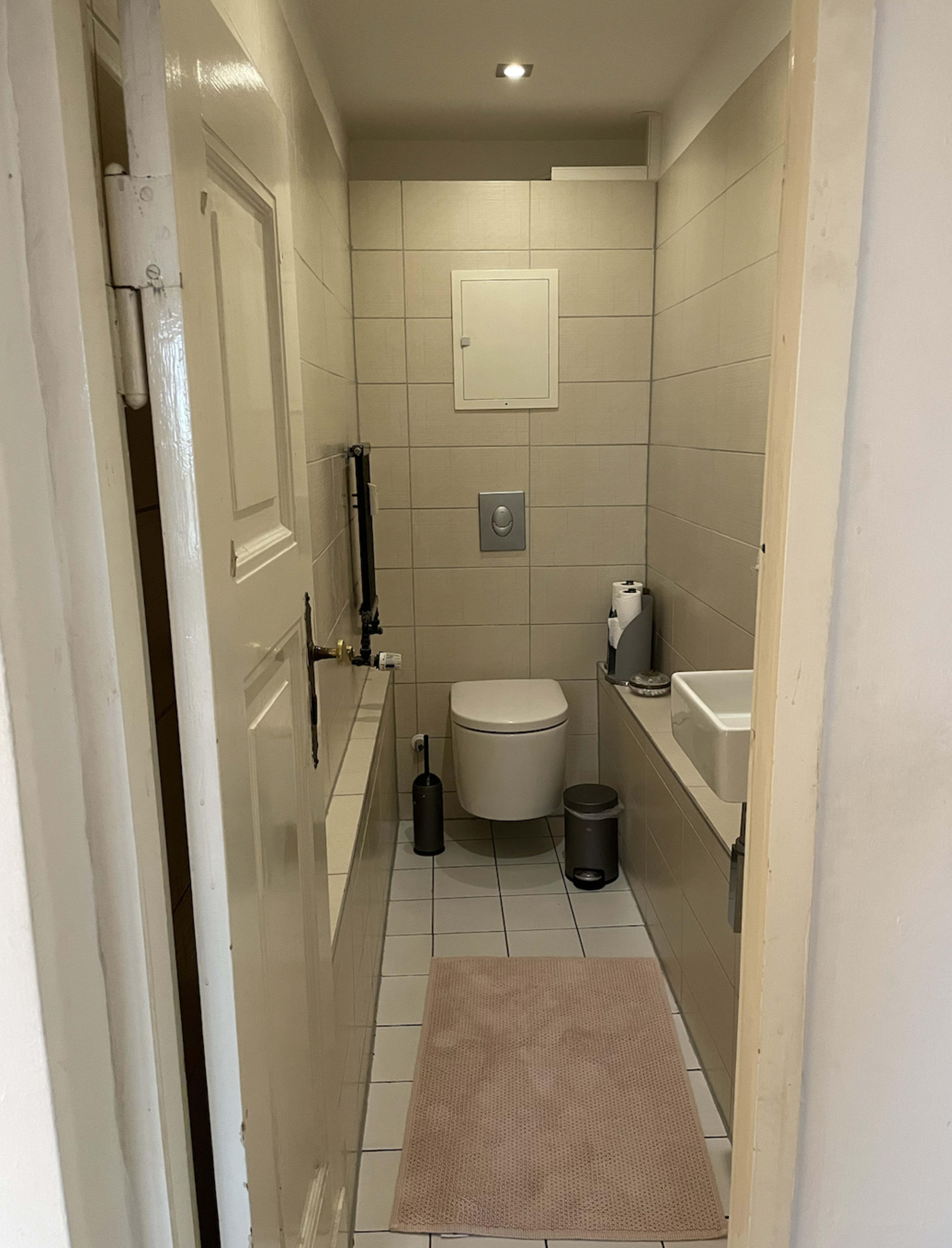 A small, tiled bathroom with a toilet, a sink, and a door leading to the hallway.