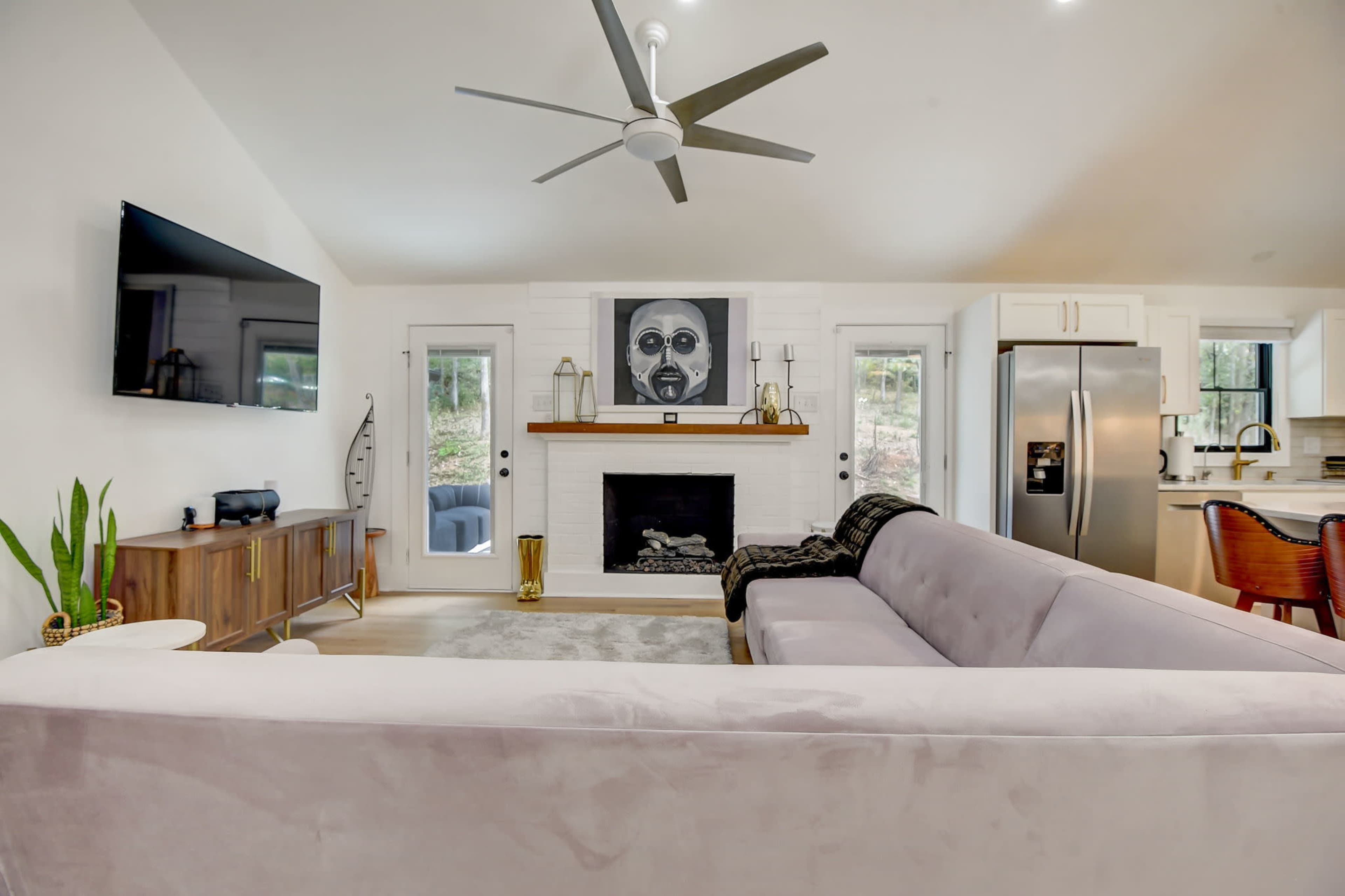 The image shows a living room featuring a light-colored sofa, a wall-mounted television, a fireplace with a decorative mantle, and a modern kitchen area in the background.