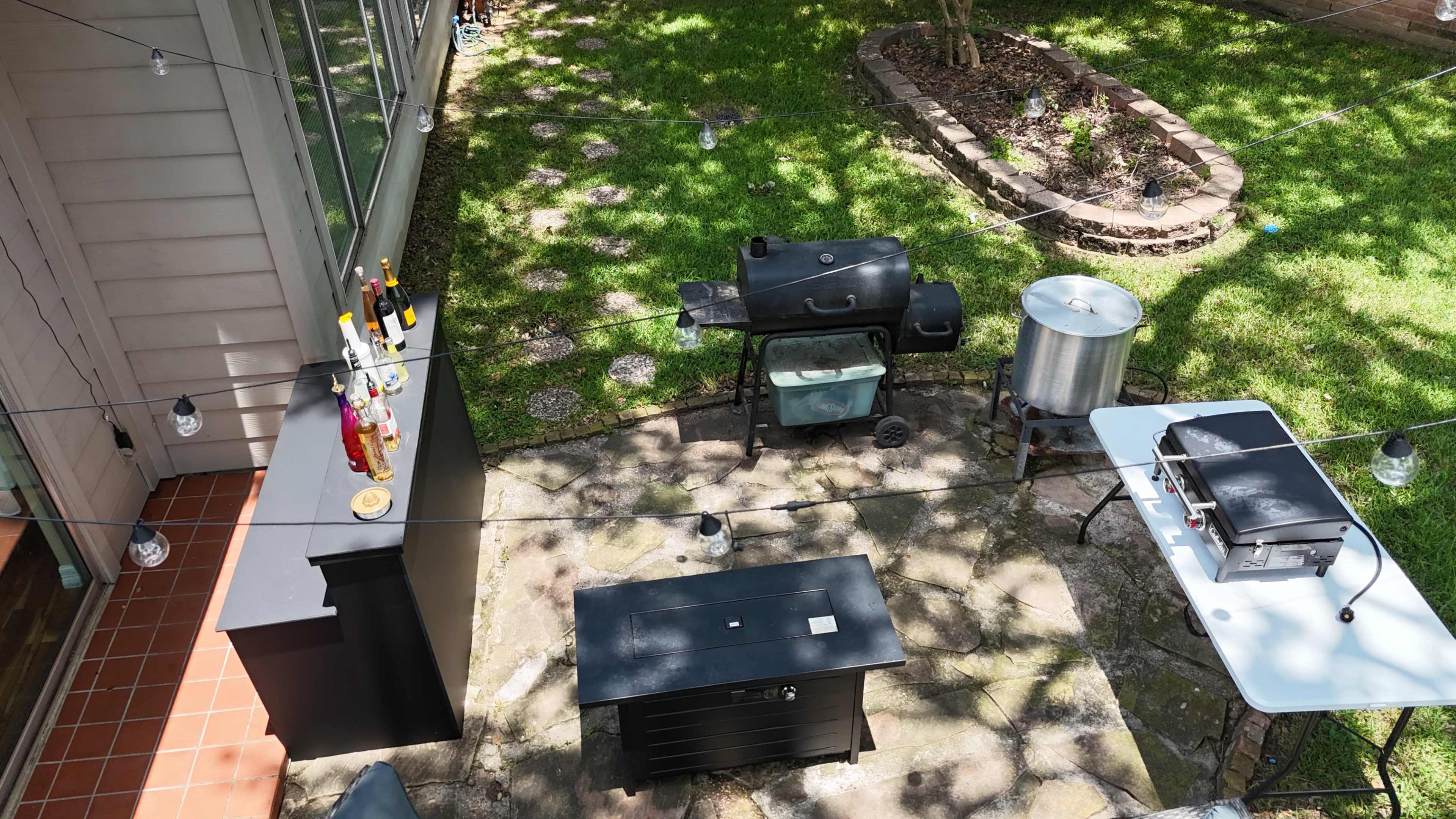 The image shows an outdoor cooking area with a variety of barbecue equipment, including a grill, a smoker, and a preparation table, surrounded by a grassy yard.