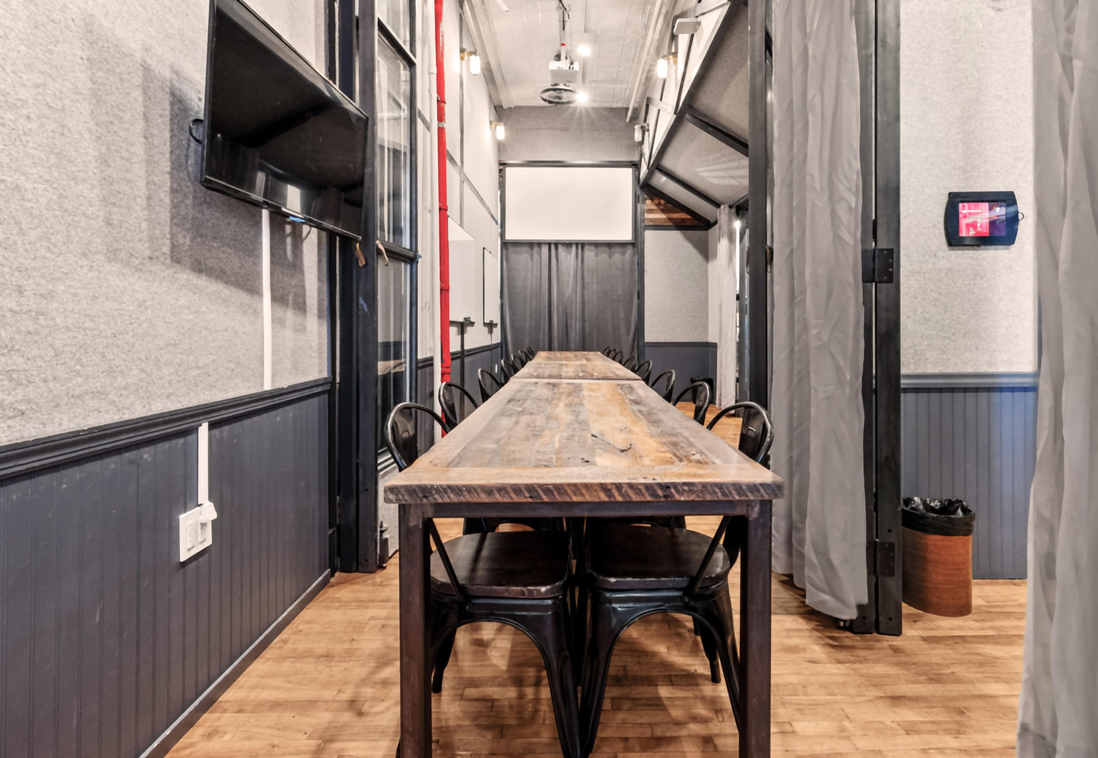 A long wooden table with black chairs is set in a narrow meeting room lined with grey walls and overhead lighting.